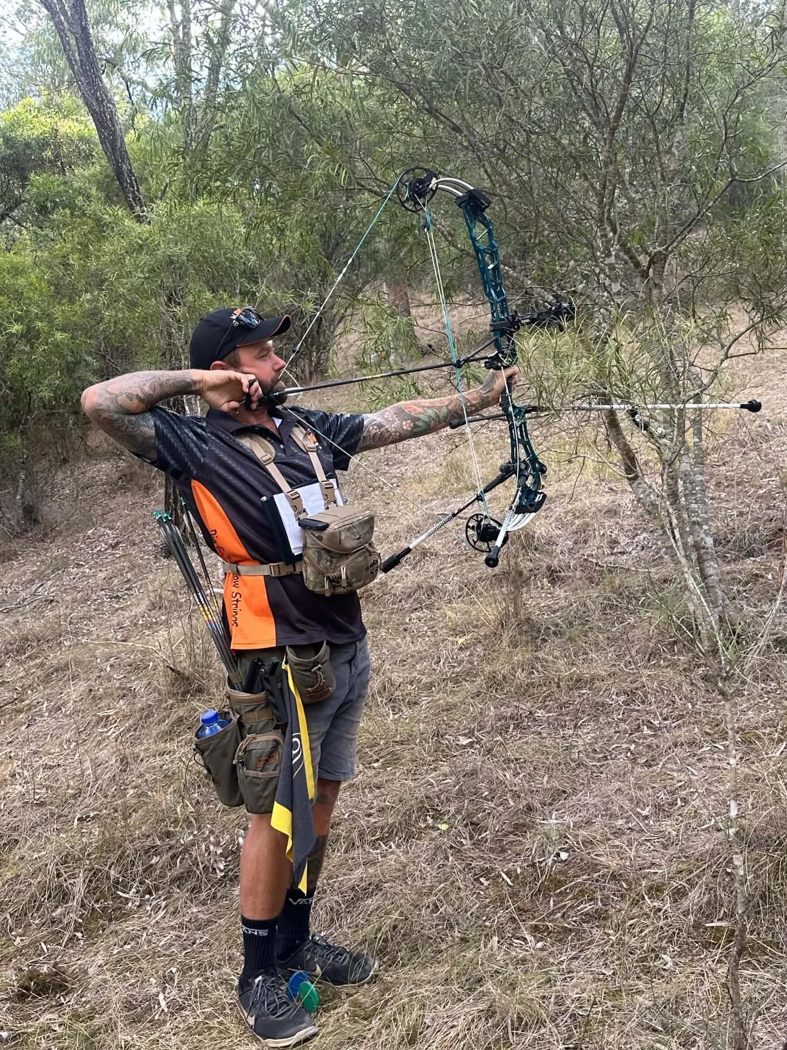 A man with tattoos and a hat practicing archery outdoors among trees, aiming a bow and arrow.