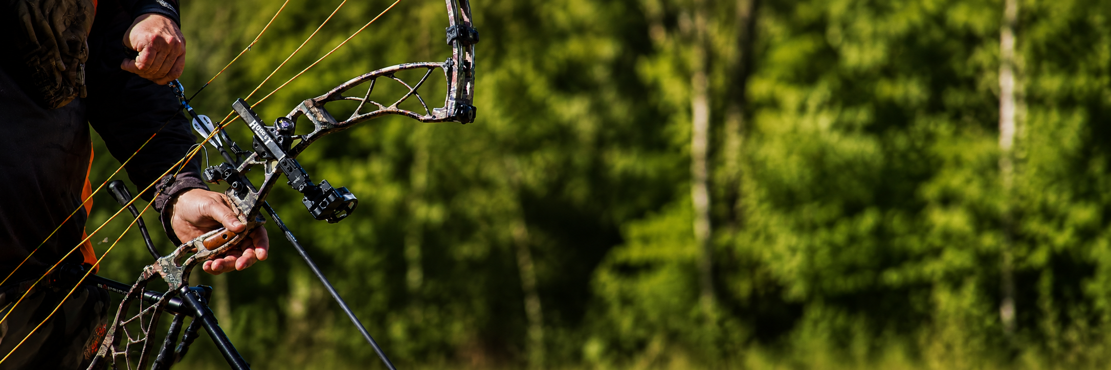 A person holding a camouflage compound bow with a quiver attached, standing outdoors in a lush green forest.