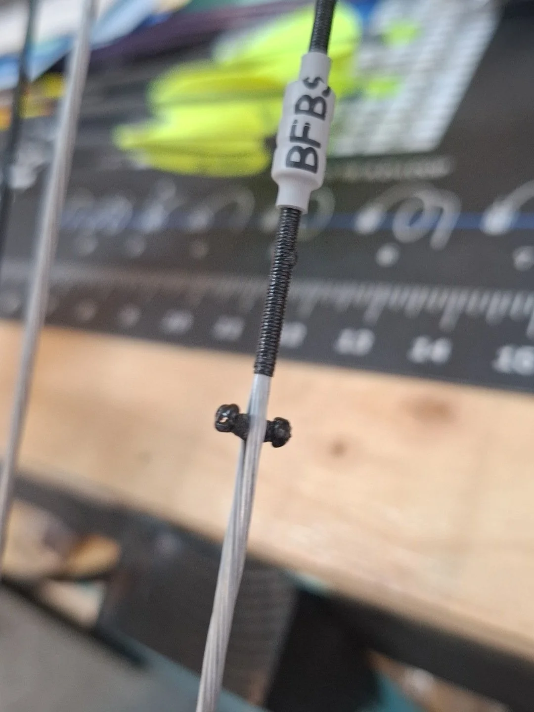 Close-up of a wire with a white label reading 'BFS' and a small black connector, likely part of a mechanical or electronic device, with a blurred background of a workbench and tools.