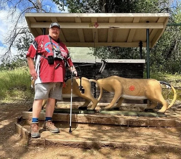A man with a red shirt and gray shorts standing next to a shooting range and a lion statue target. The lion statue has a red bulls-eye target on its side and is part of a shooting practice setup. The scene is outdoors with trees in the background.