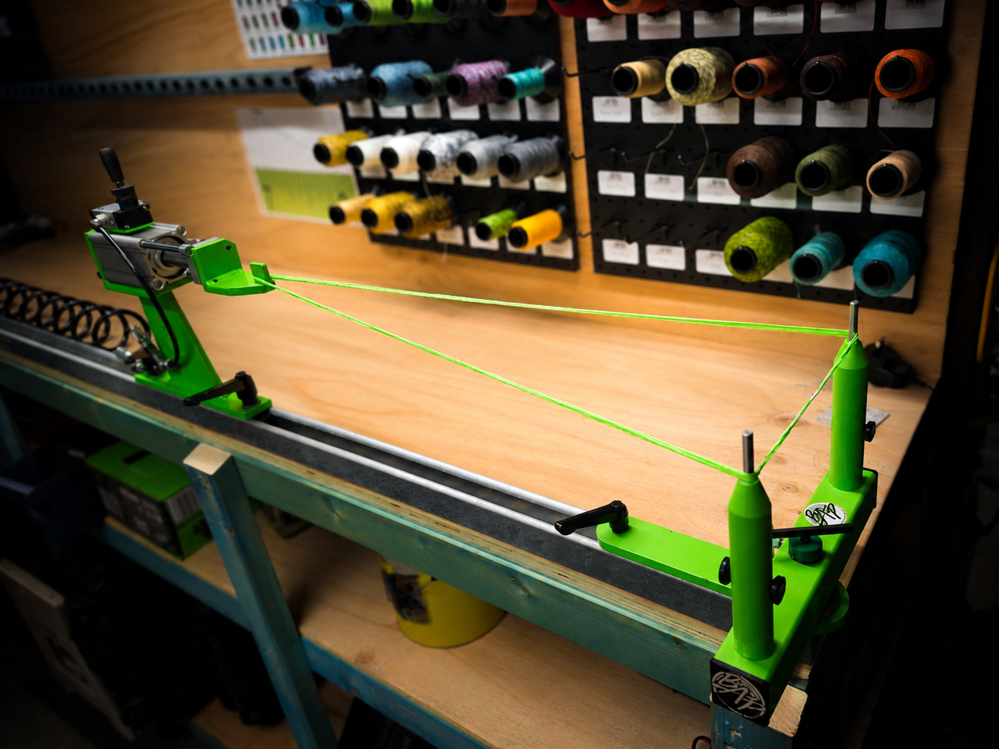 A green string tension device on a workbench in a workshop with spools of colorful thread on a pegboard in the background.