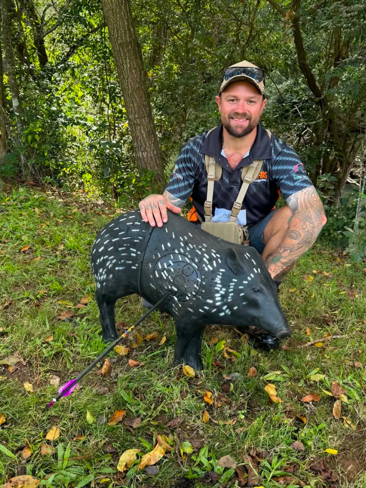 A man with tattoos, wearing a camouflage cap, sunglasses on his head, and a dark sporting shirt, kneels outdoors in a wooded area while holding a black boar sculpture decorated with white dots and paint. An arrow with colorful fletching lies on the grass nearby.