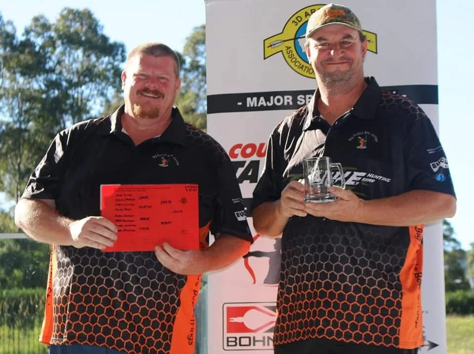 Two men standing outdoors smiling, one holding a red certificate and the other holding a glass award, in front of a banner with logos and text.