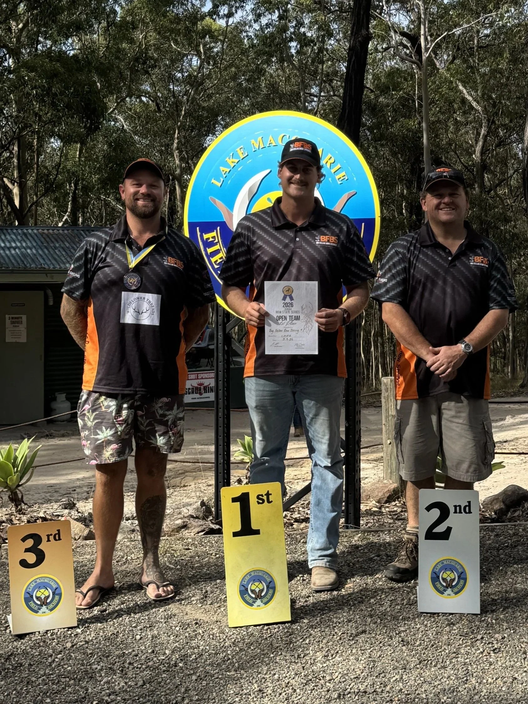 Three men standing outdoors behind a winners' podium at a fishing competition. The man in the middle is holding a certificate and standing in first place, flanked by second and third place winners. The background has trees and a blue and yellow sign that says 'Lake Macquarie'.
