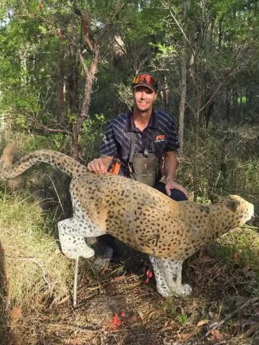 Man kneeling in a forested area with a large carved wooden or fake animal resembling a leopard.