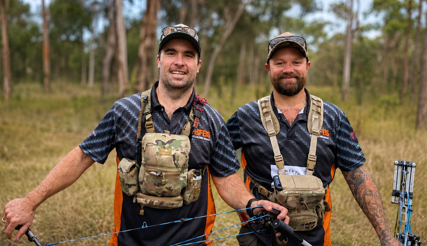 Two men standing outdoors in a forest, wearing black and orange shirts with camouflage gear, smiling, holding archery equipment.
