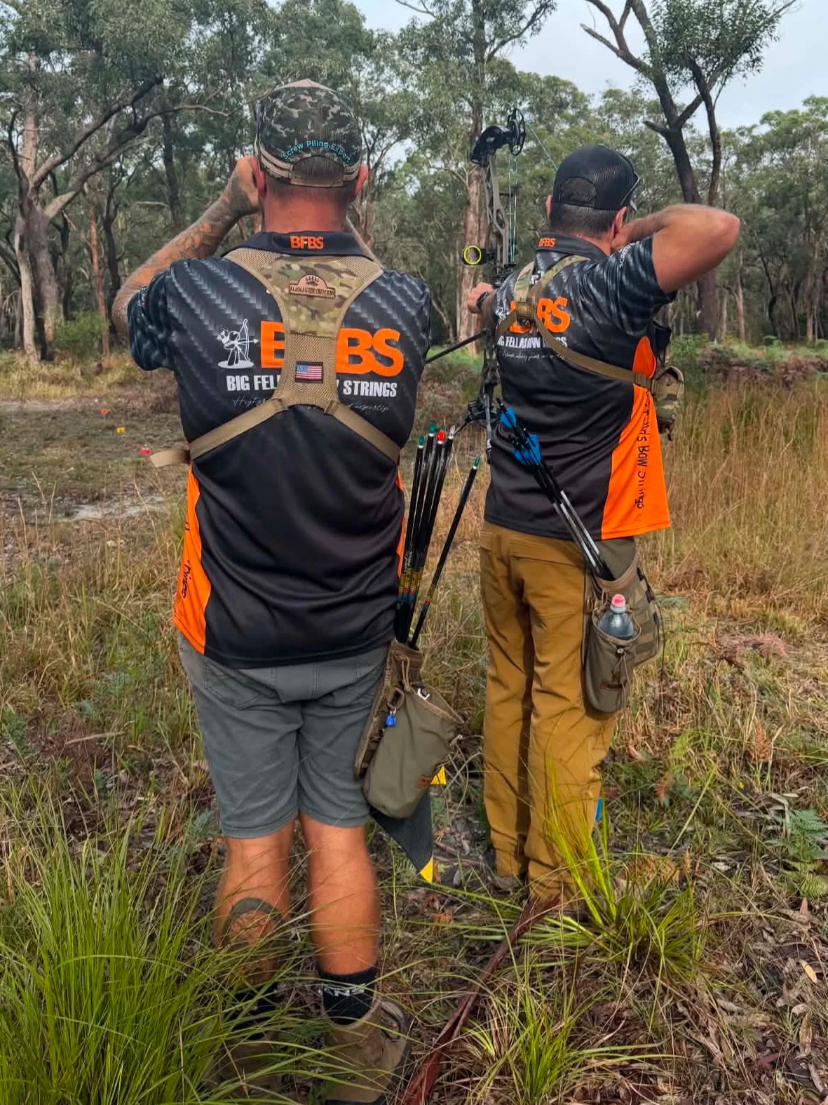 Two men archery shooting in a wooded clearing, wearing matching black and orange shirts with archery equipment, standing among tall grass and trees.