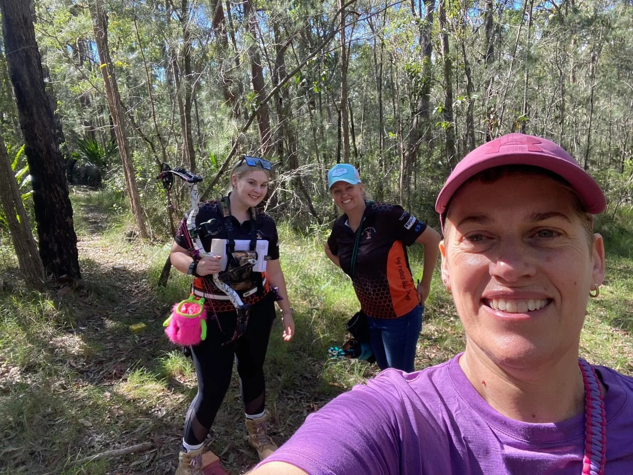 Three women and part of a fourth woman taking a selfie, standing in a wooded area during the daytime. They are dressed in casual outdoor clothing, with two wearing hats and one with sunglasses on her head.