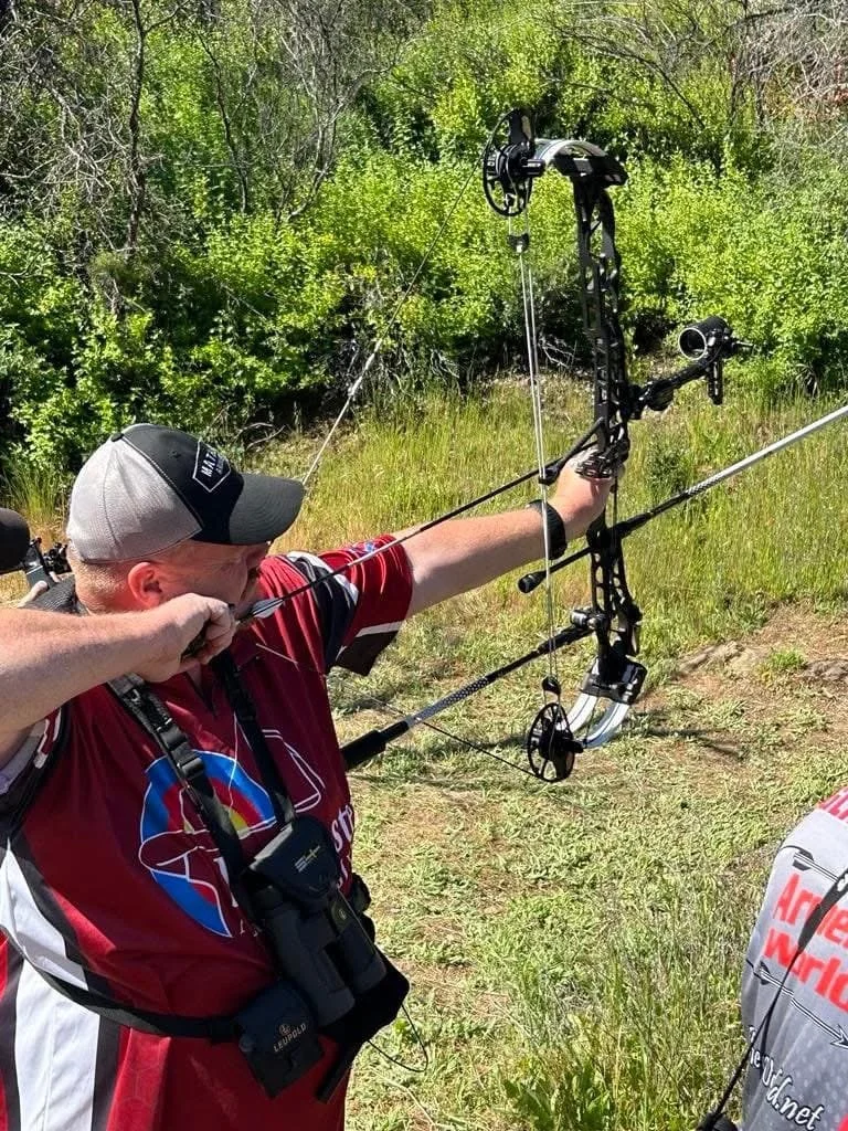 Man practicing archery outdoors in a grassy area with green trees in the background, aiming a compound bow.