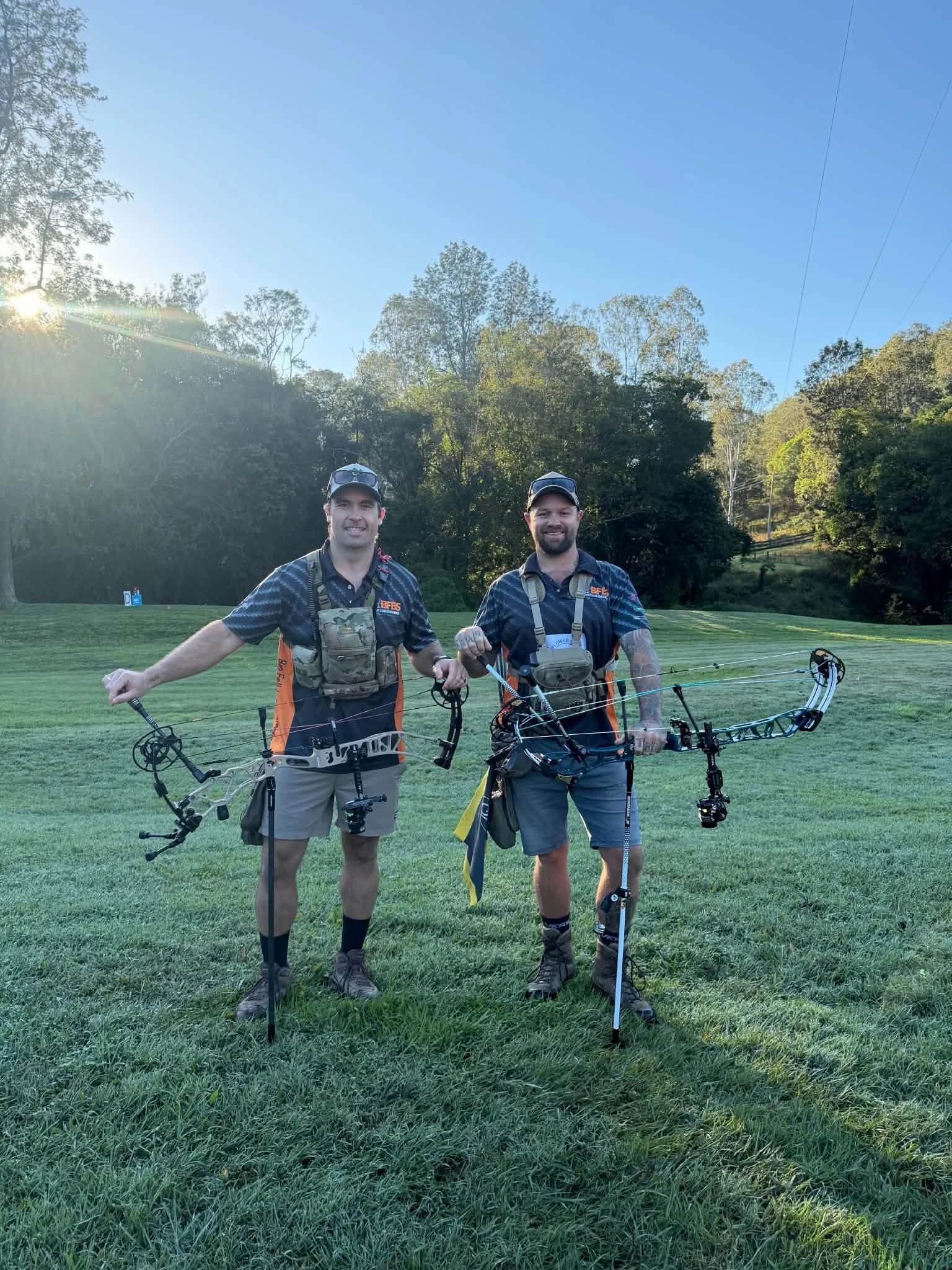 Two men standing on a grassy field holding bows, smiling, with trees and a bright blue sky in the background