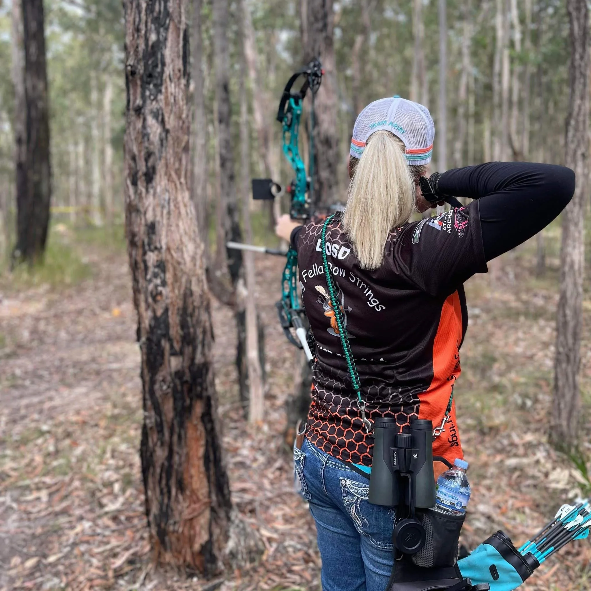 A woman with long blonde hair tied in a ponytail, wearing a white cap with turquoise and orange accents, a black and orange athletic jacket, and blue jeans, is aiming a blue bow and arrow in a wooded area with tall trees and fallen leaves. She is standing with her back to the camera, holding the bowstring and aiming at a target out of view. She has a water bottle hanging from her waist and a quiver with arrows on her hip.