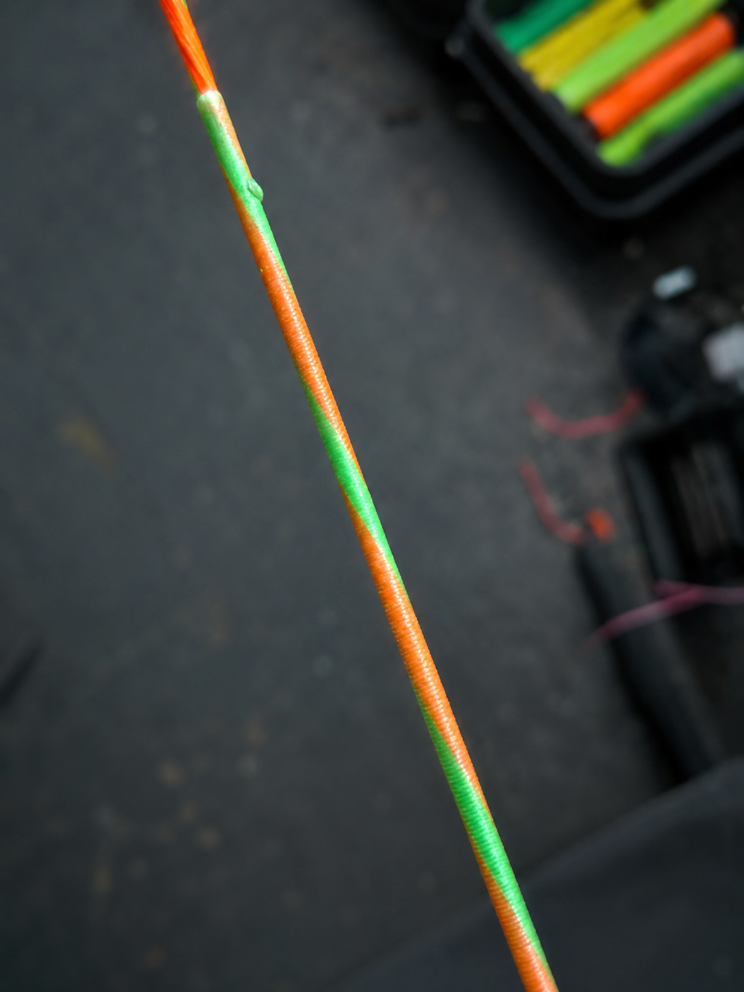 Close-up of a fishing rod wrapped with orange and green tape, with fishing tackle box in the background.