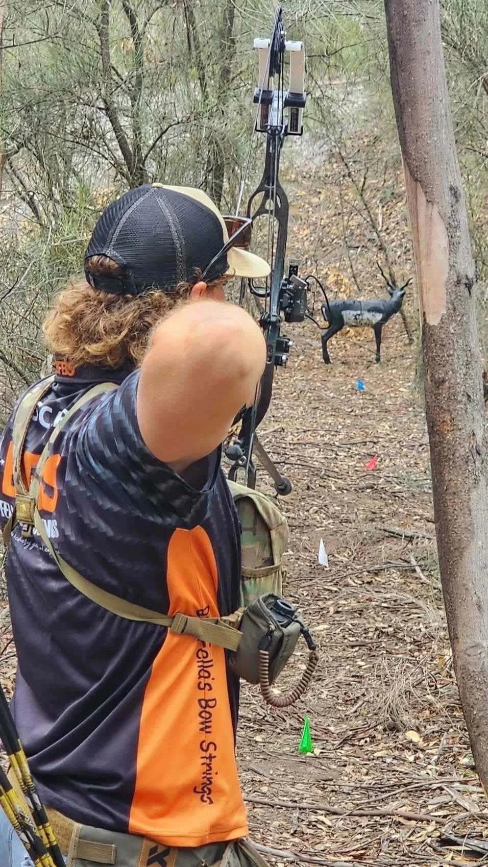 A person with curly hair and a black and yellow cap aims a bow and arrow at a paper deer target in a wooded area during archery practice.