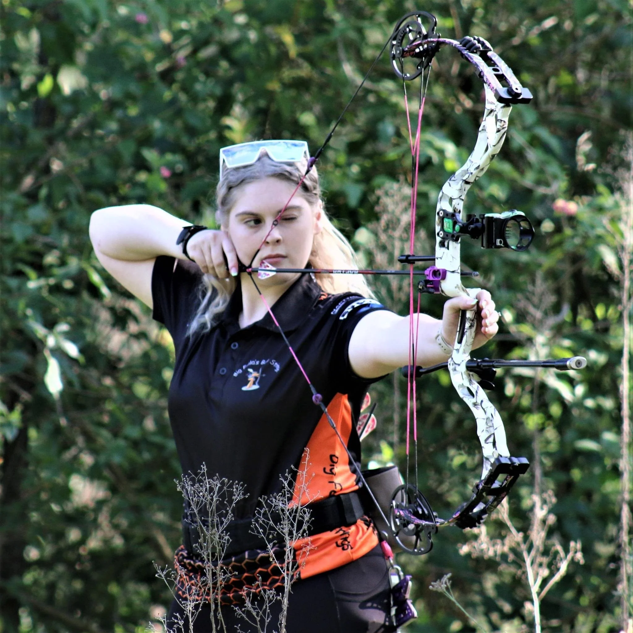 A young girl with glasses aims a compound bow and arrow in a wooded area.