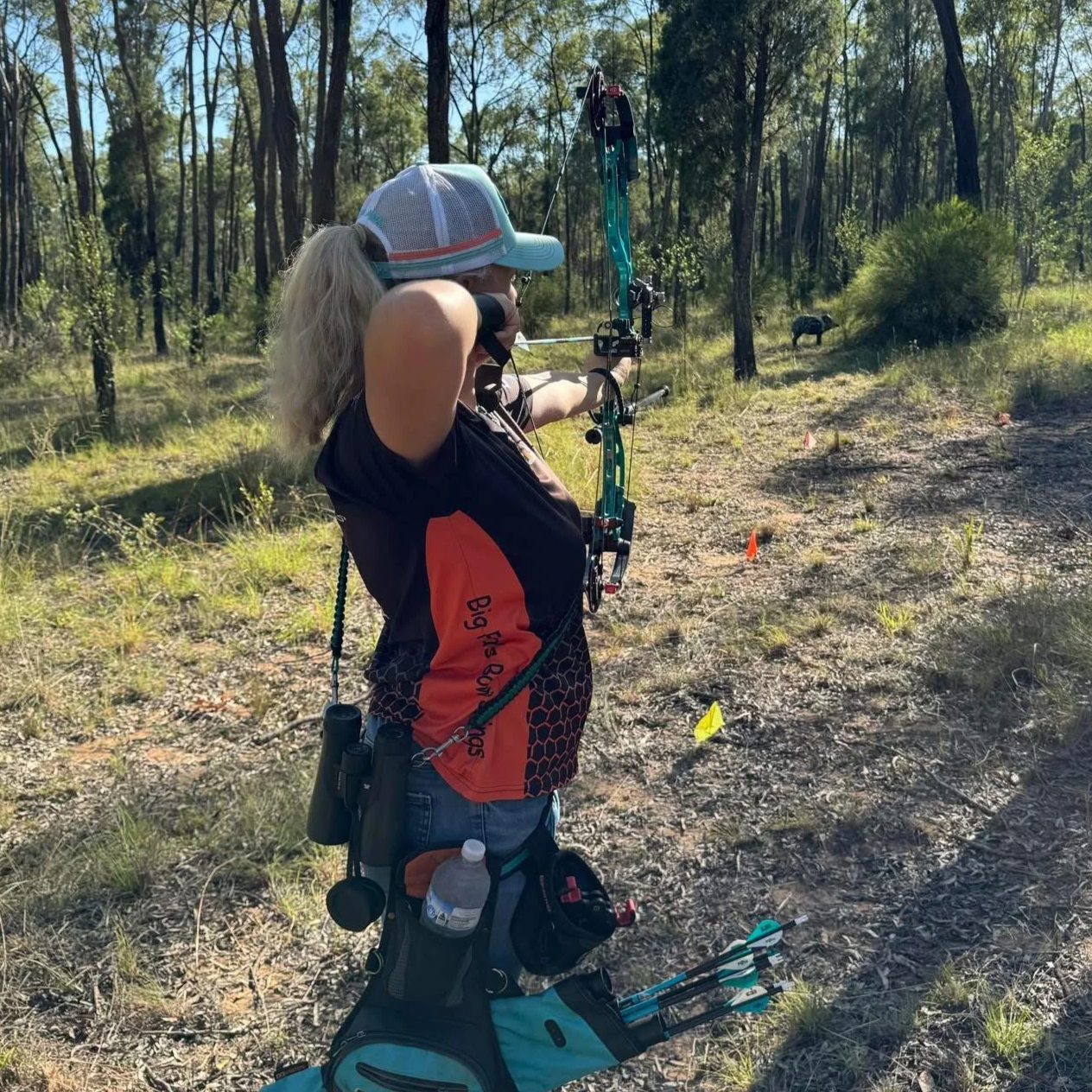 A woman with an arrow nocked in her bow, aiming in a forested area during daylight. The woman has blonde hair, a white cap, and is wearing sports gear. She is preparing to shoot, with her right arm extended and left hand holding the bow. A bag with more arrows is seen on the ground nearby.