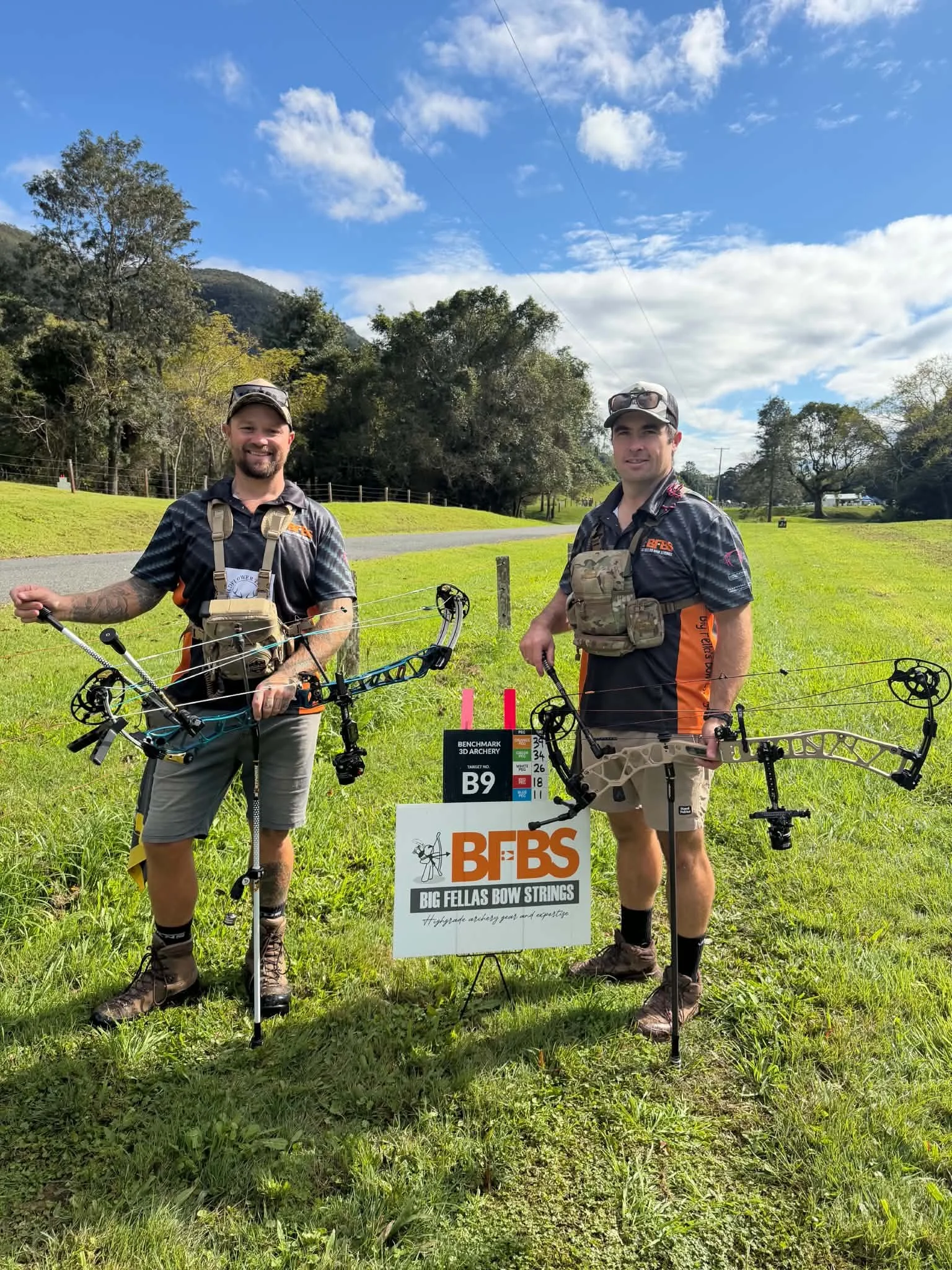 Two men standing outdoors holding bows, with a sign that reads "BFB Big Fellas Bow Strings." They are dressed in outdoor gear and sunglasses, smiling at the camera, with green grass, trees, and a partly cloudy sky in the background.