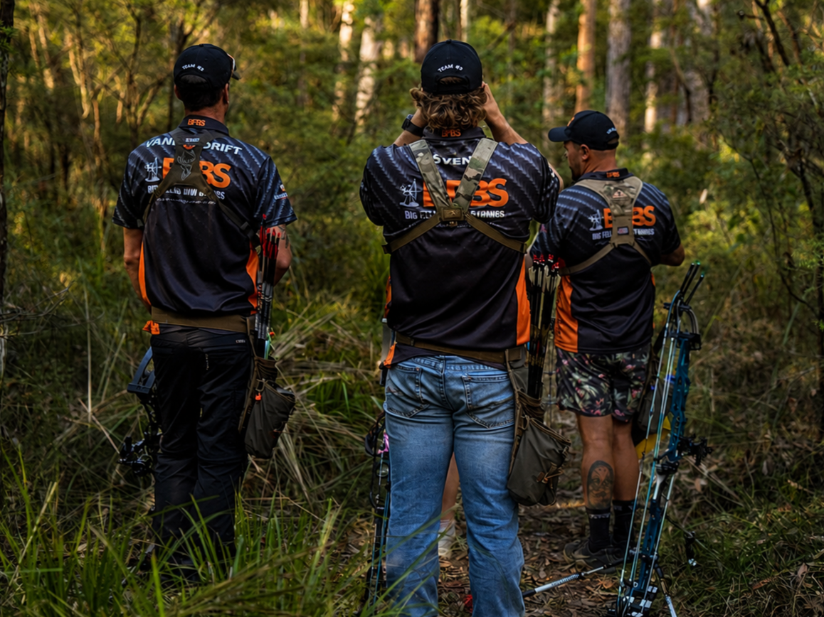 Three people stand in a wooded area, wearing archery shirts and gear, with one person holding a bow, likely preparing for or engaging in an archery activity.