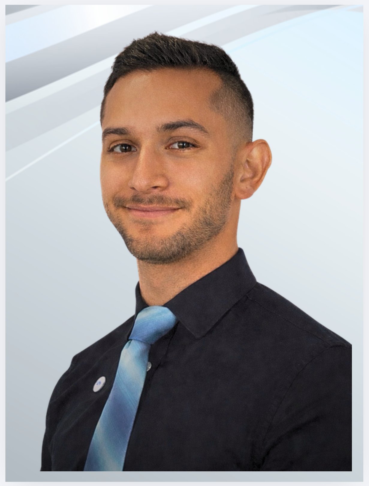 A young man with short dark hair and a beard, wearing a black shirt and a blue tie with light and dark shades, smiling at the camera against a light blue and white gradient background.