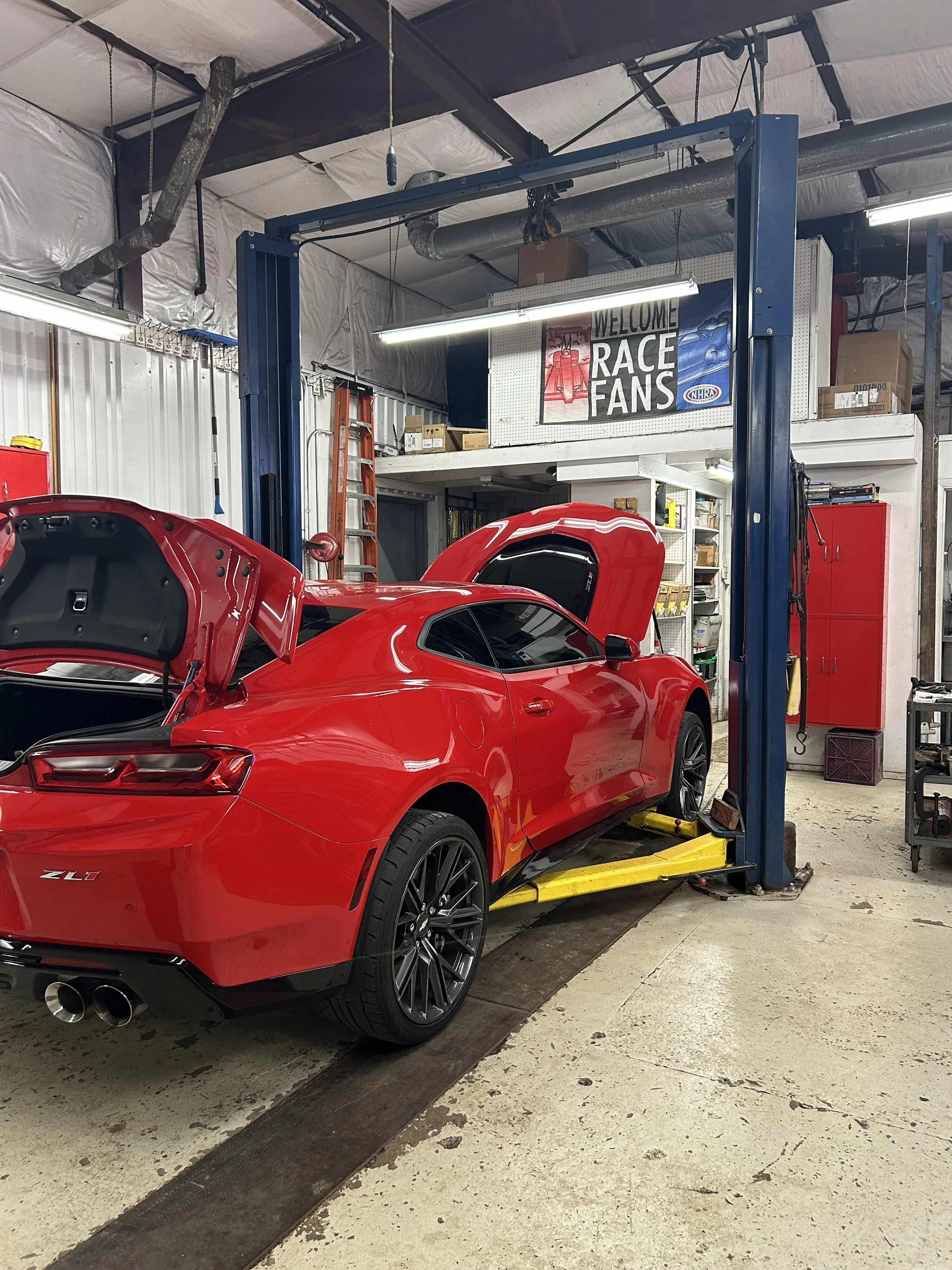 Red Chevrolet Camaro ZL1 sports car in an auto repair garage on a hydraulic lift with raised hood and trunk open.