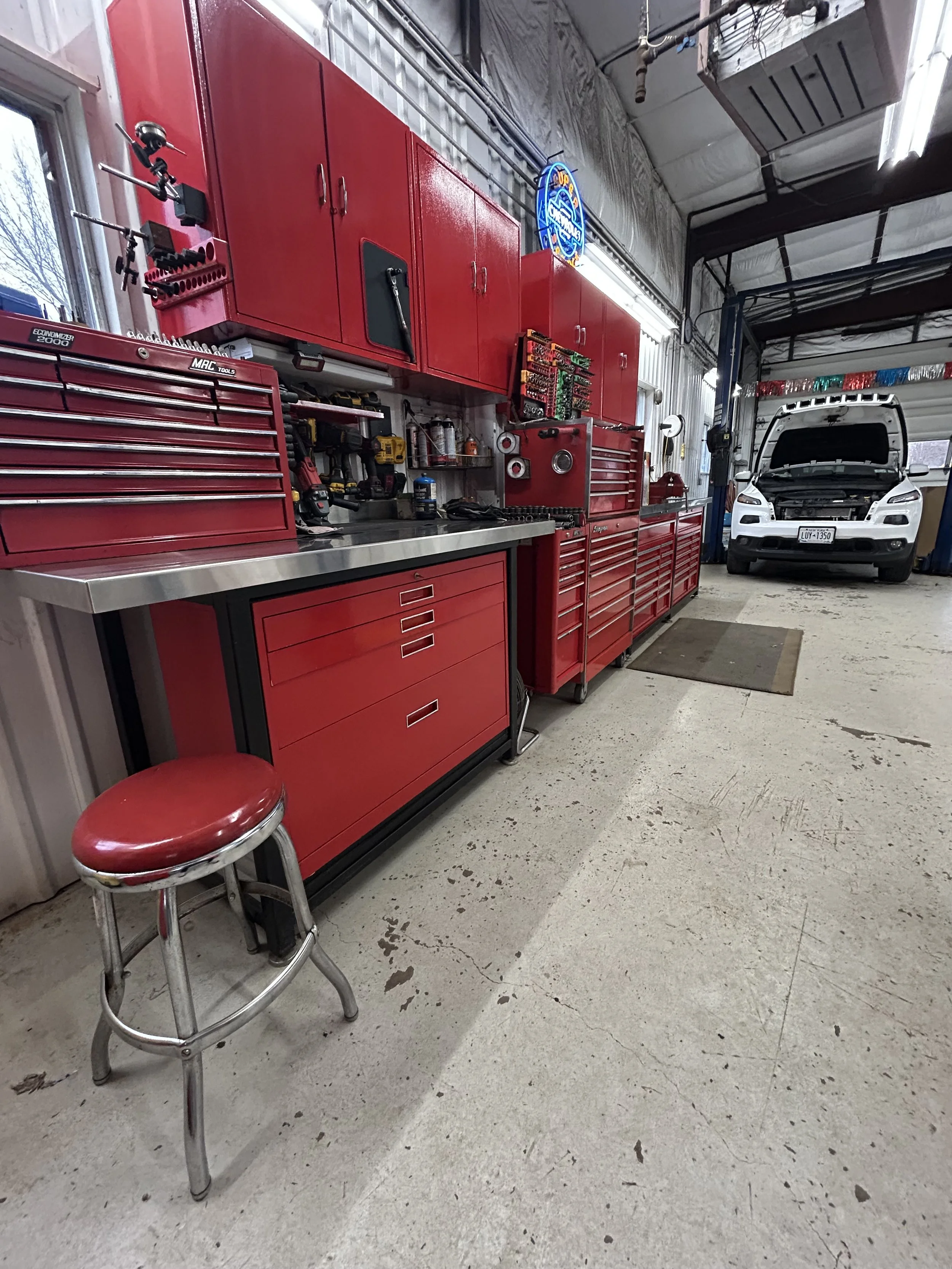 Inside a garage with a red tool cabinet, workbench, and storage units. A stool with a red seat in front of the work area. A car with an open hood parked inside the garage, with a concrete floor.