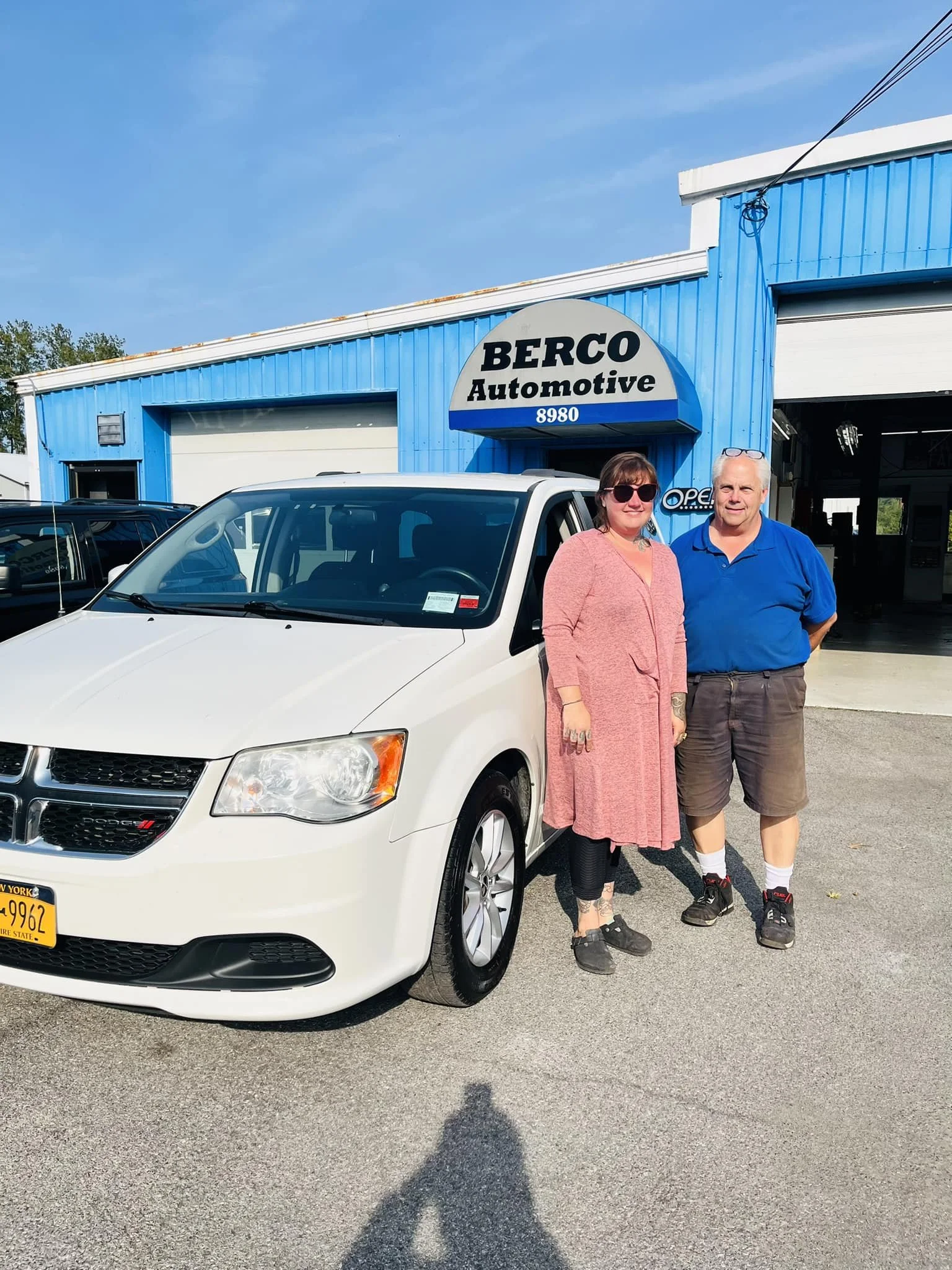 A couple standing next to a white Dodge car in front of a blue building with a sign that reads 'BERCO Automotive'.