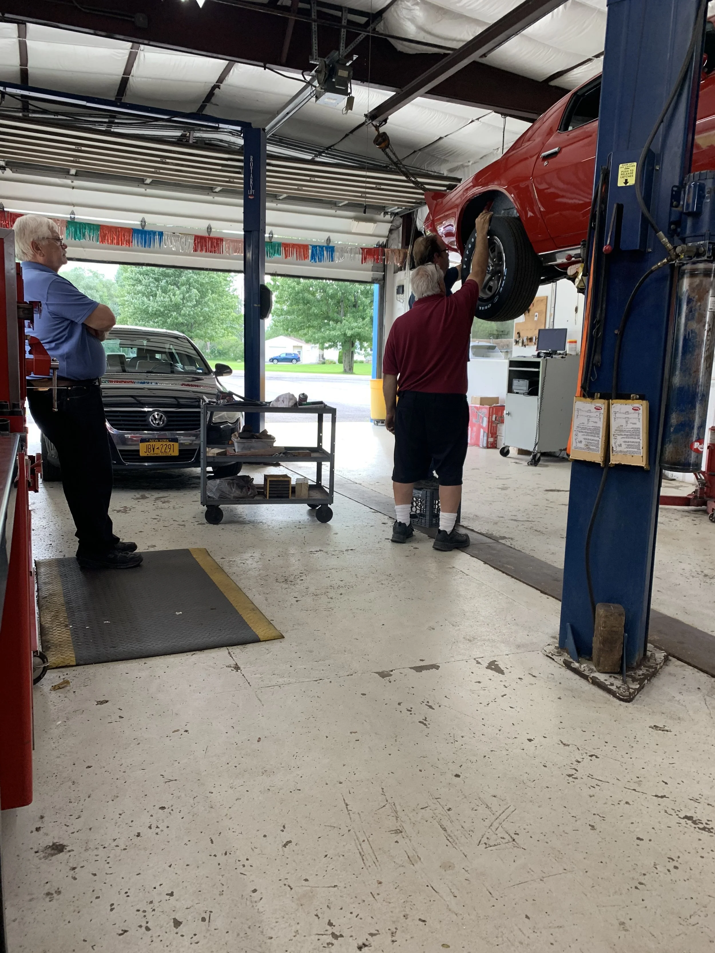 Three men in a garage, with one standing on a crate, working on a red car lifted by a mechanic’s hoist, and another man watching. A black Volkswagen vehicle is parked inside, with a residential area visible outside.