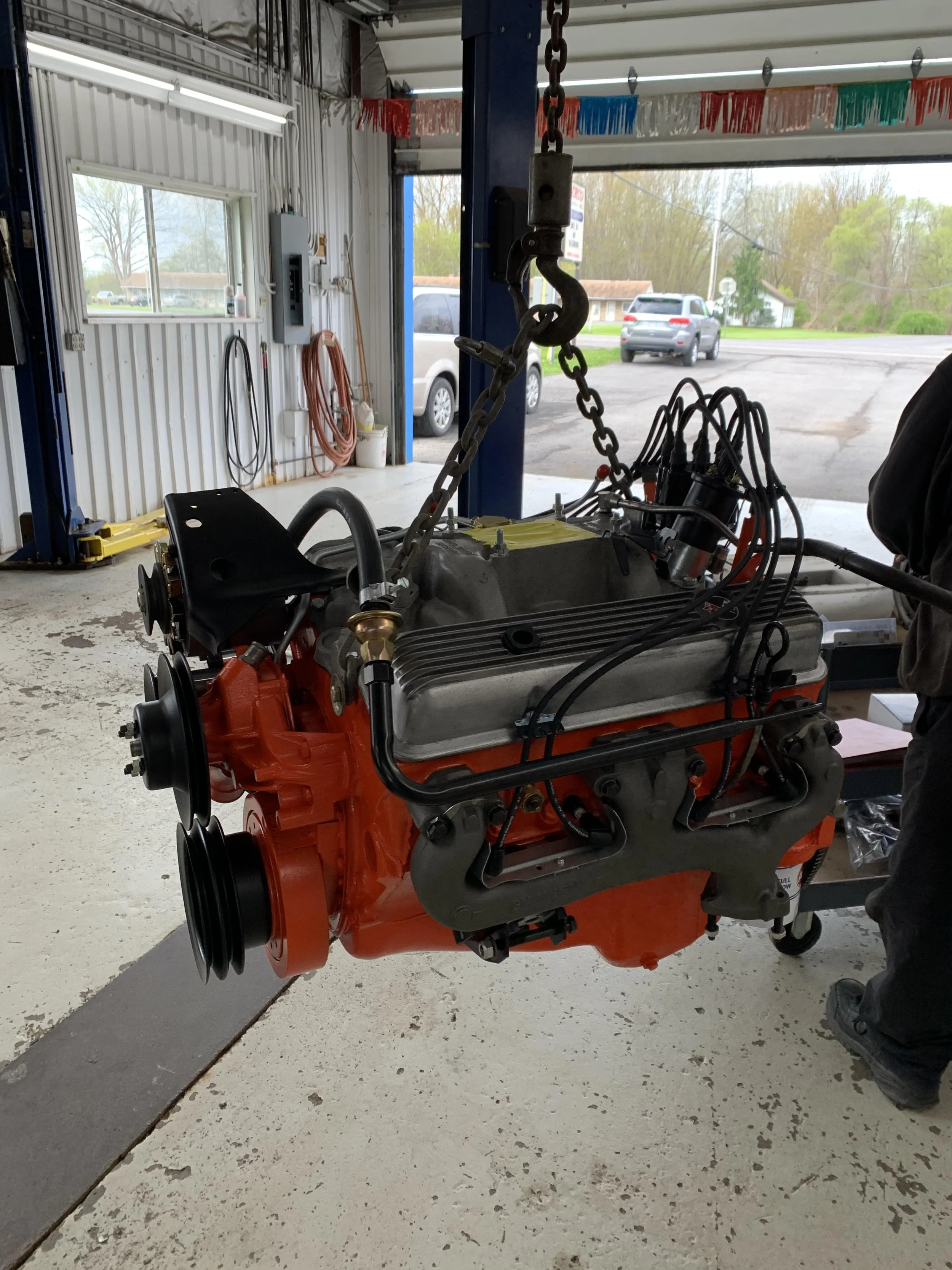 An orange engine with black cables and silver components is suspended in a garage, with cars visible through the open door outside.