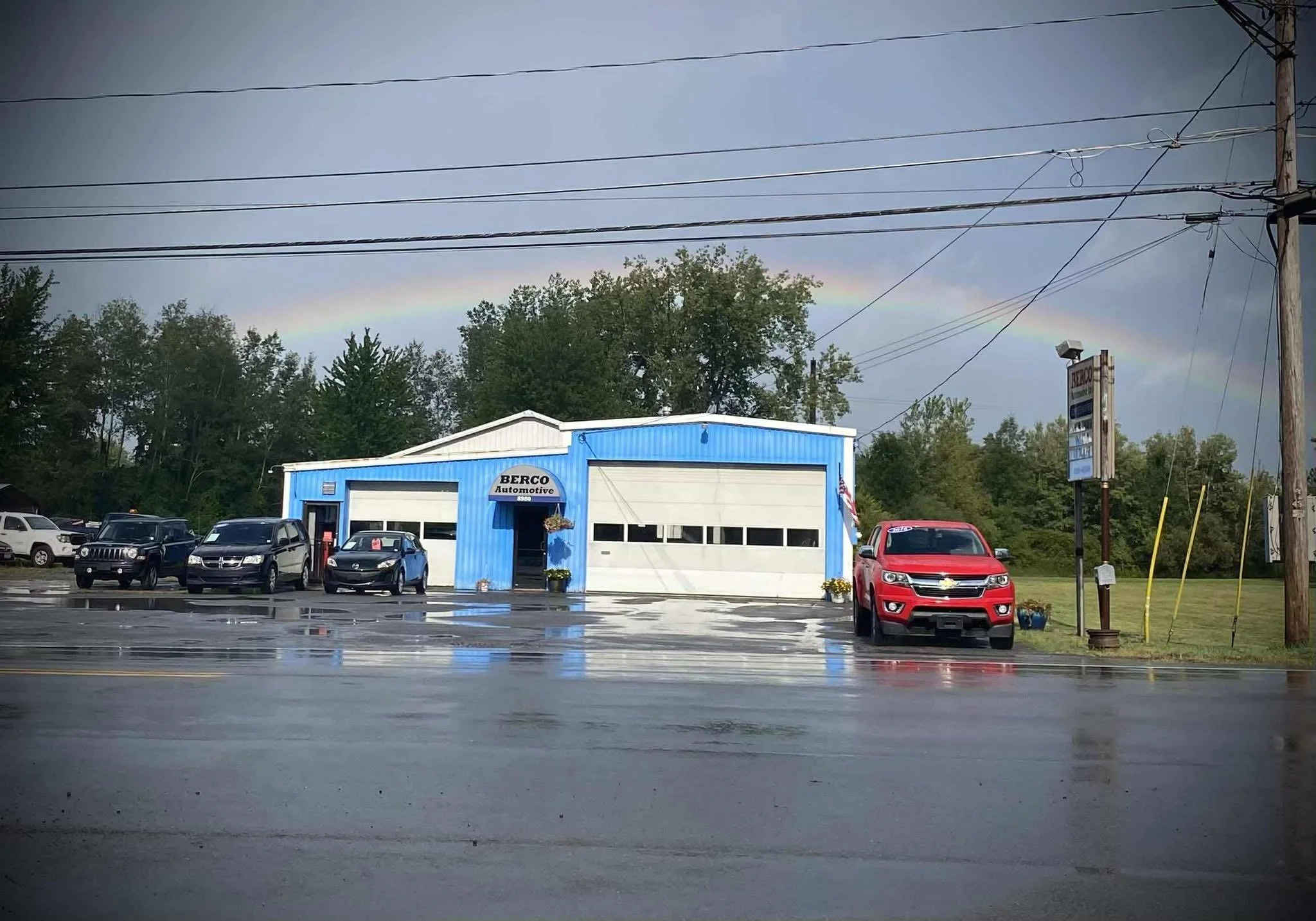 Car dealership with a blue building, several parked cars, a rainbow in the cloudy sky, and a wet road in front.