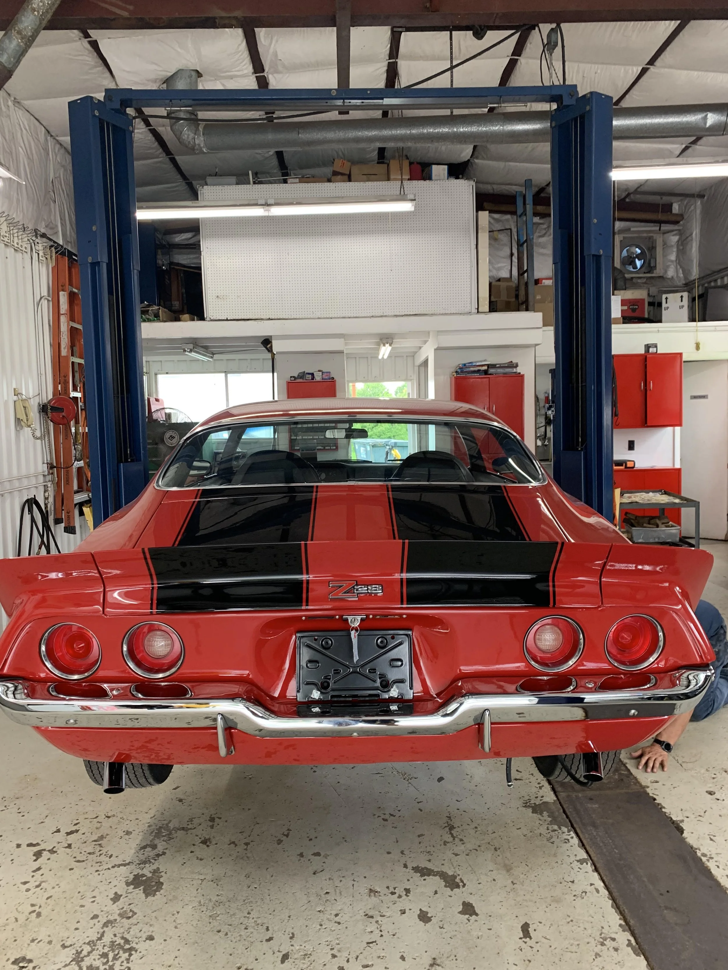 Red Chevrolet Corvette Z28 with black racing stripes, parked in a garage with red storage cabinets, a pegboard with tools, and a workshop area. The car is on a lift with a person partially visible underneath.