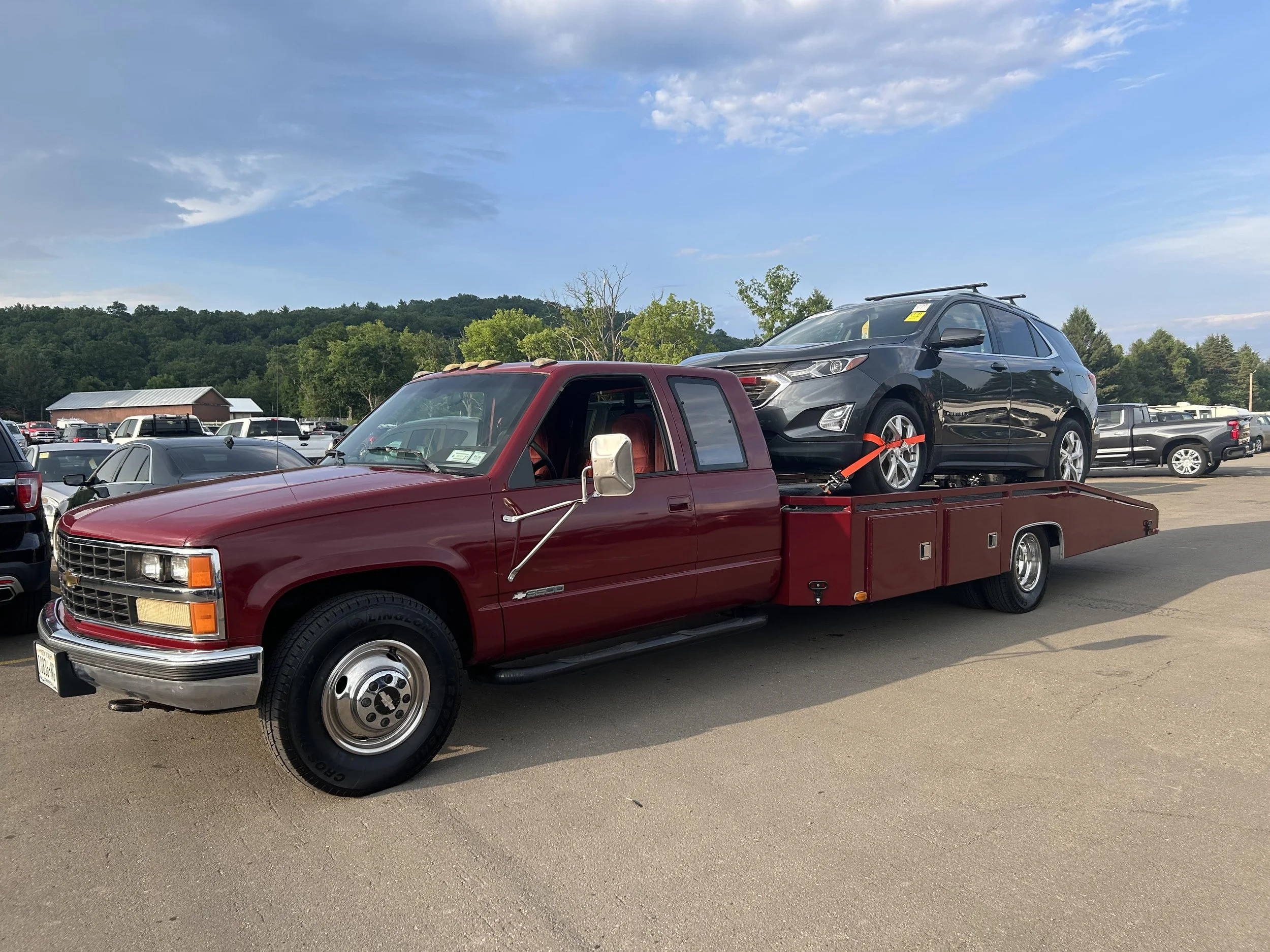 A red flatbed tow truck carrying a black sedan in a parking lot with other vehicles and trees in the background under a partly cloudy sky.