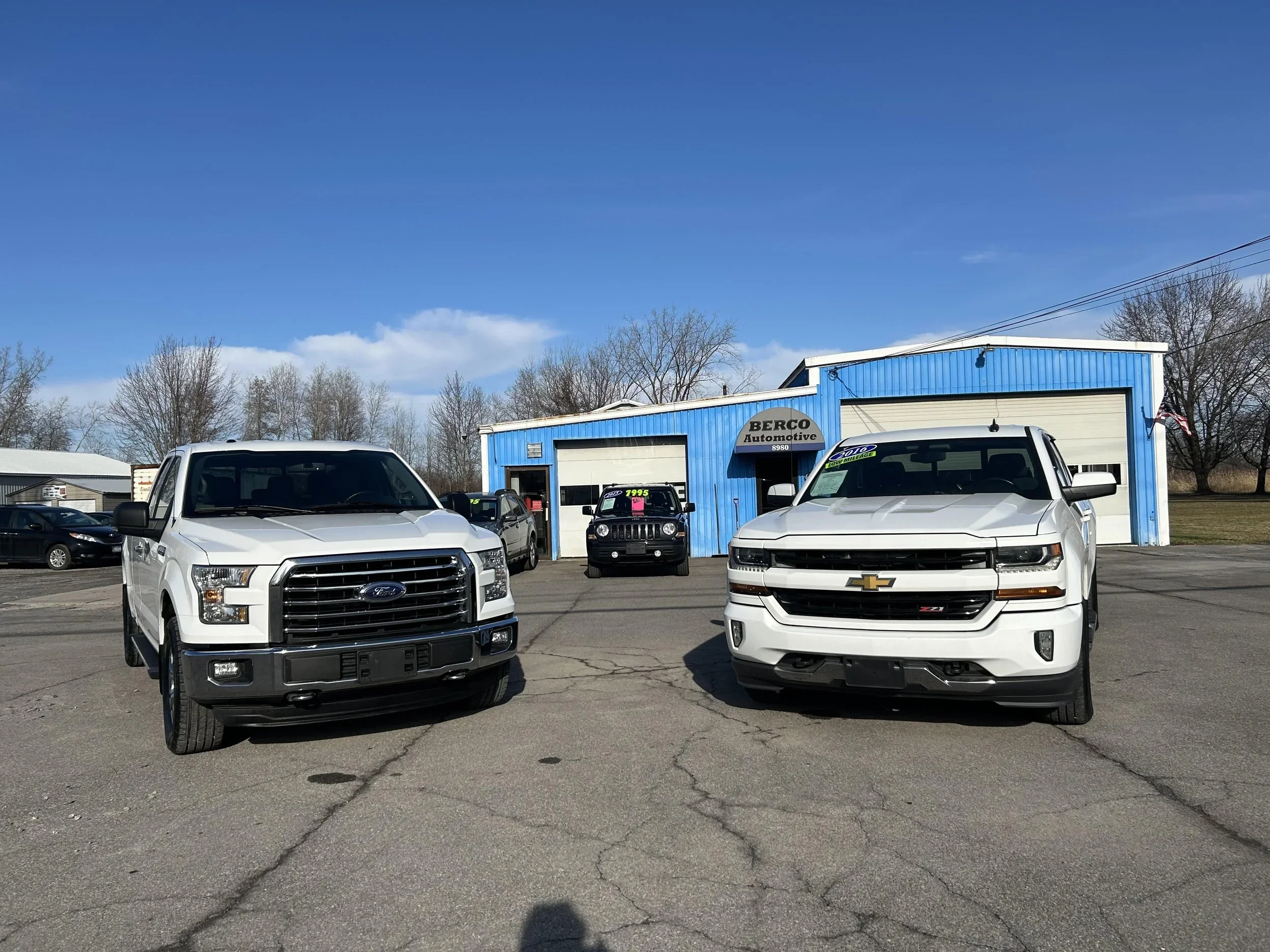 A white Ford pickup truck and a white Chevrolet SUV parked in front of a blue auto dealership building with a sign reading "Berco Automotive." Several other cars are visible in the background under a partly cloudy sky.