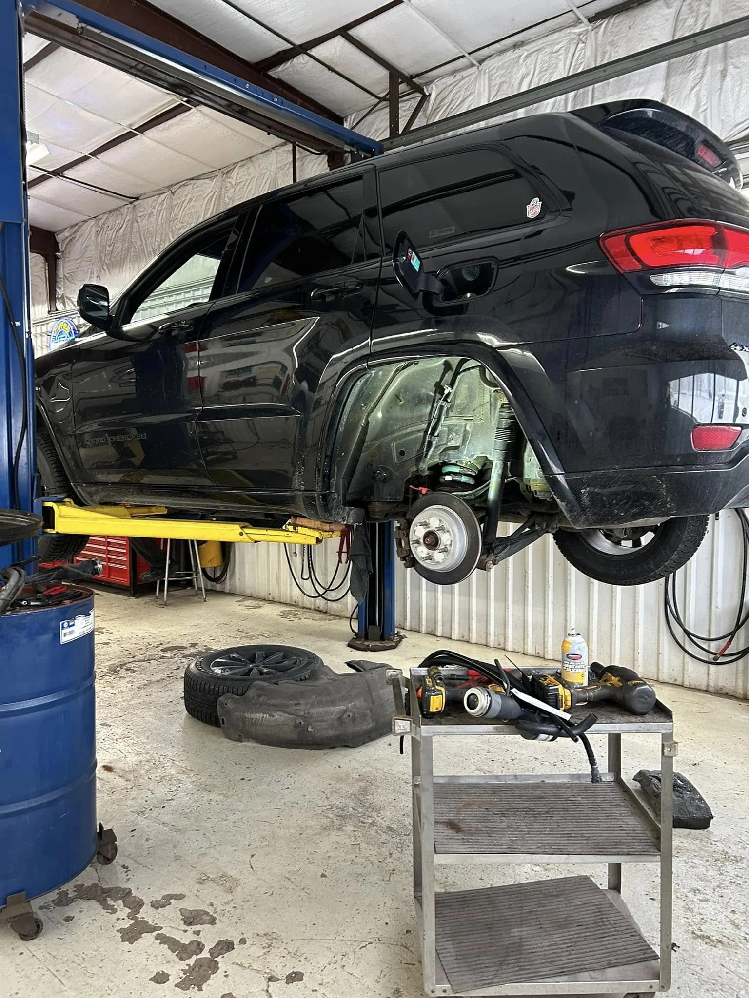 Black SUV elevated on a hydraulic lift with the front wheels removed, exposing the brake assembly. The garage workspace includes tools and an engine part on a cart, an extra tire, and a blue barrel.