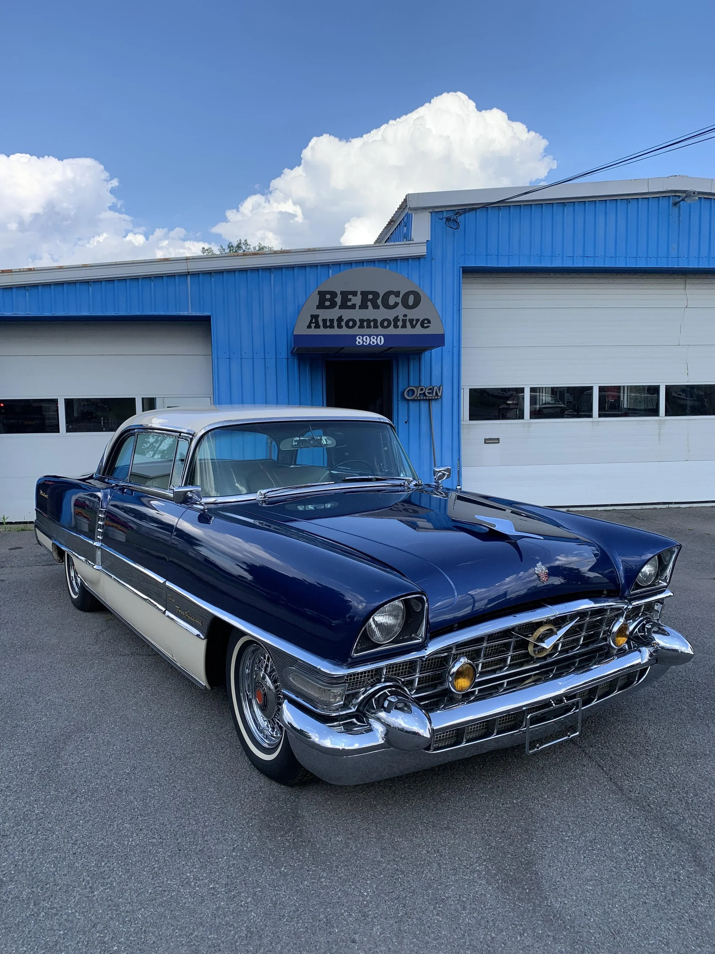 A classic blue and white vintage car parked outside a blue automotive shop named BERCO Automotive, with a partly cloudy sky in the background.