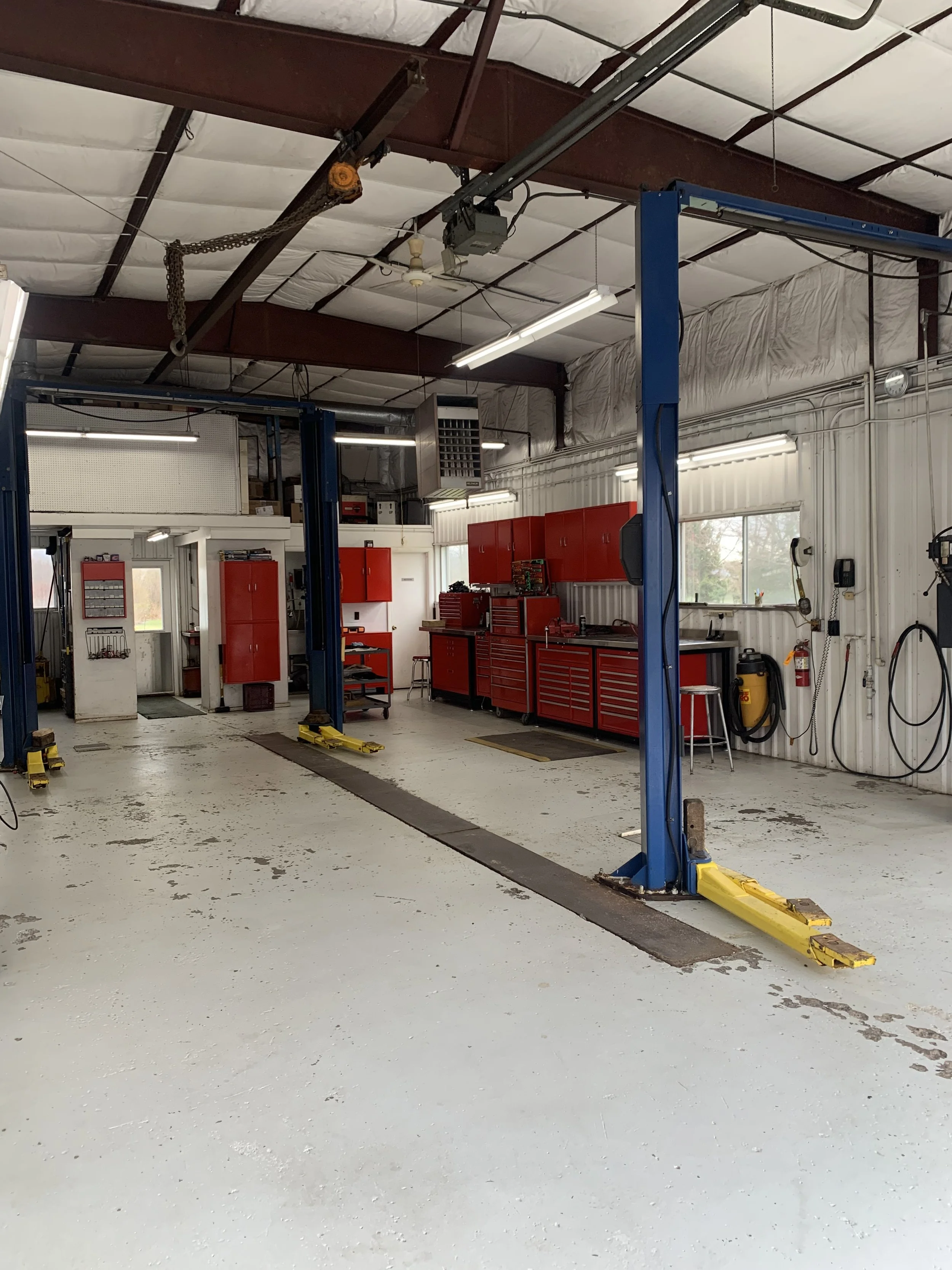 Empty automotive repair shop with red tool cabinets, workbenches, and a vehicle lift in the center.