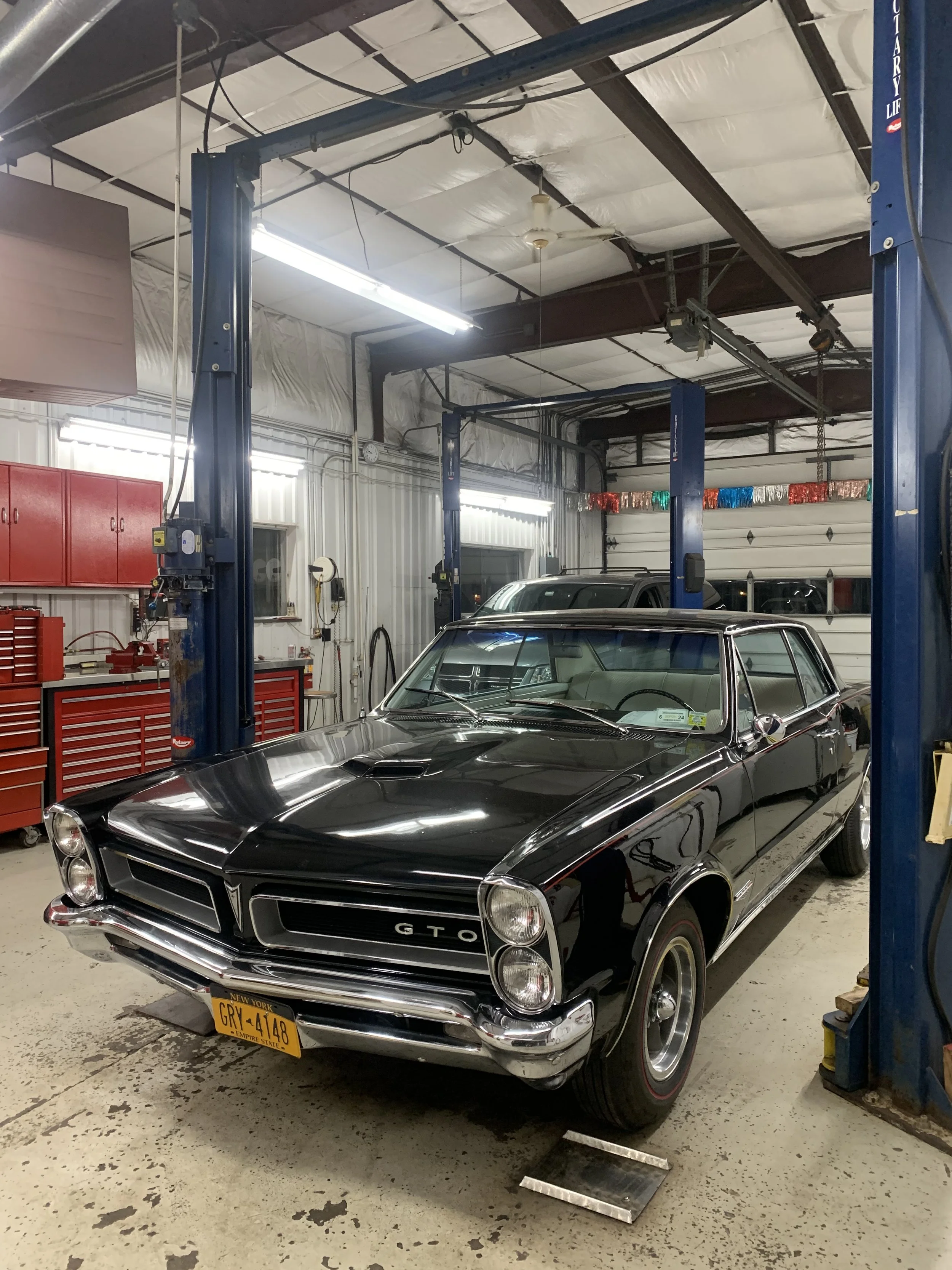 A black vintage Pontiac GTO muscle car inside a well-lit garage or auto repair shop with red tool cabinets and shelves in the background.