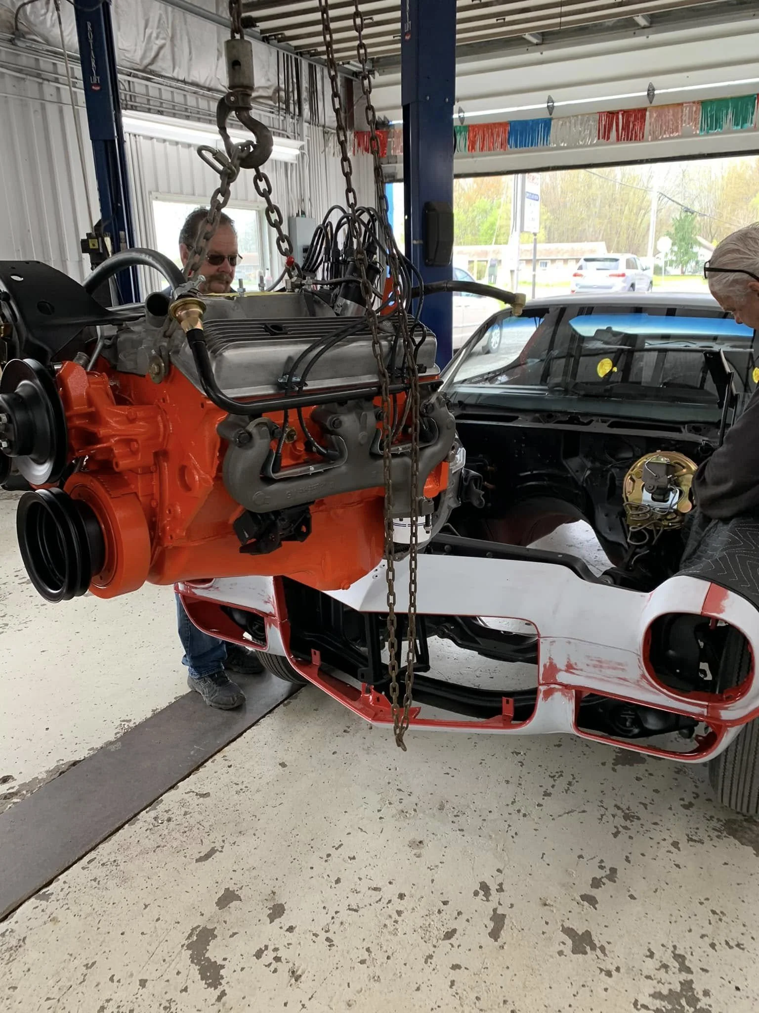 A car under construction in a garage, with two workers installing a large orange engine into the front of the car.