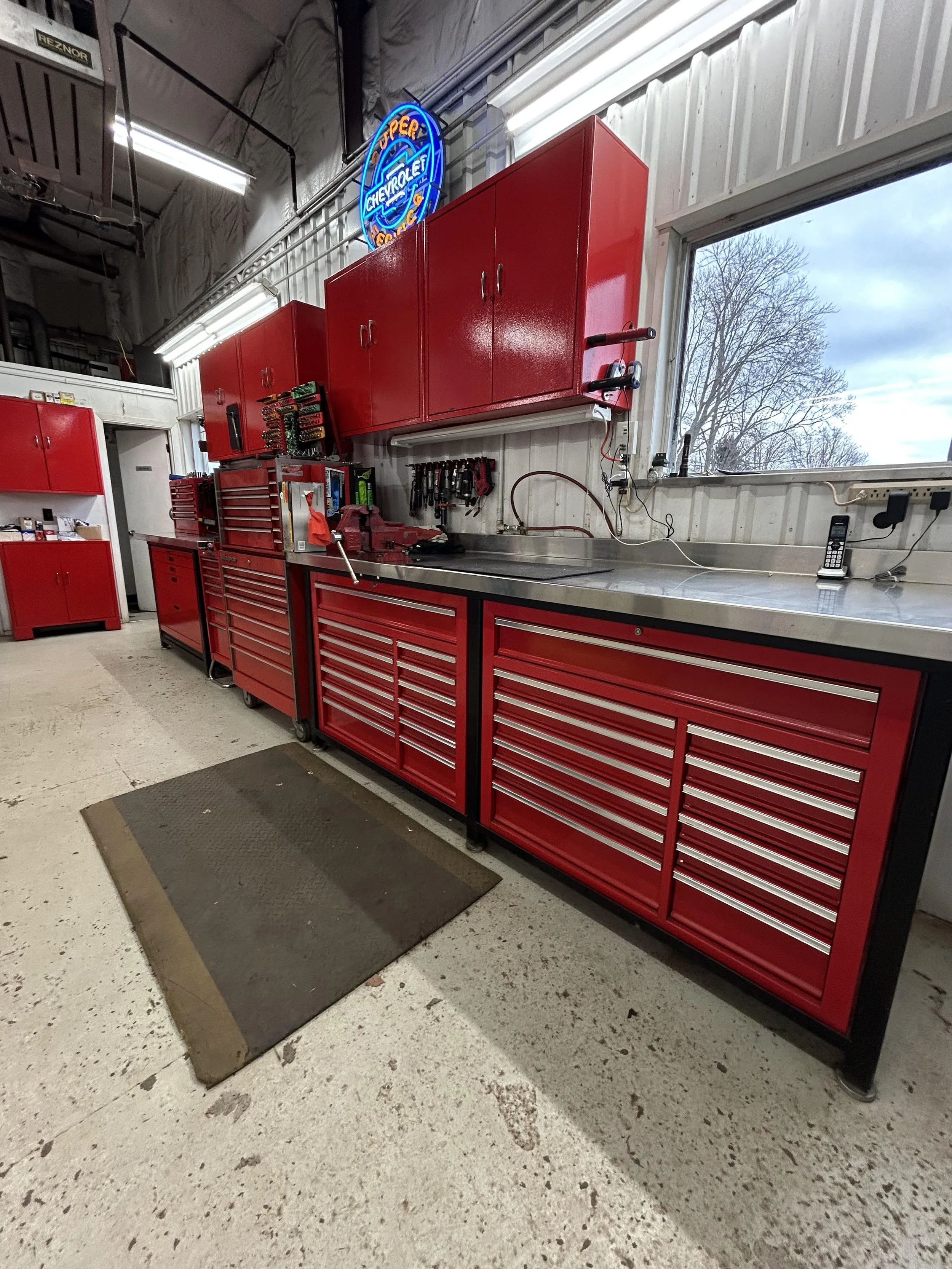 Red tool cabinets with drawers and shelves in a workshop, a rubber mat on the floor, a window showing cloudy sky and trees outside, and a neon sign that says 'Chevrolet' in blue and orange, with other words partially visible.