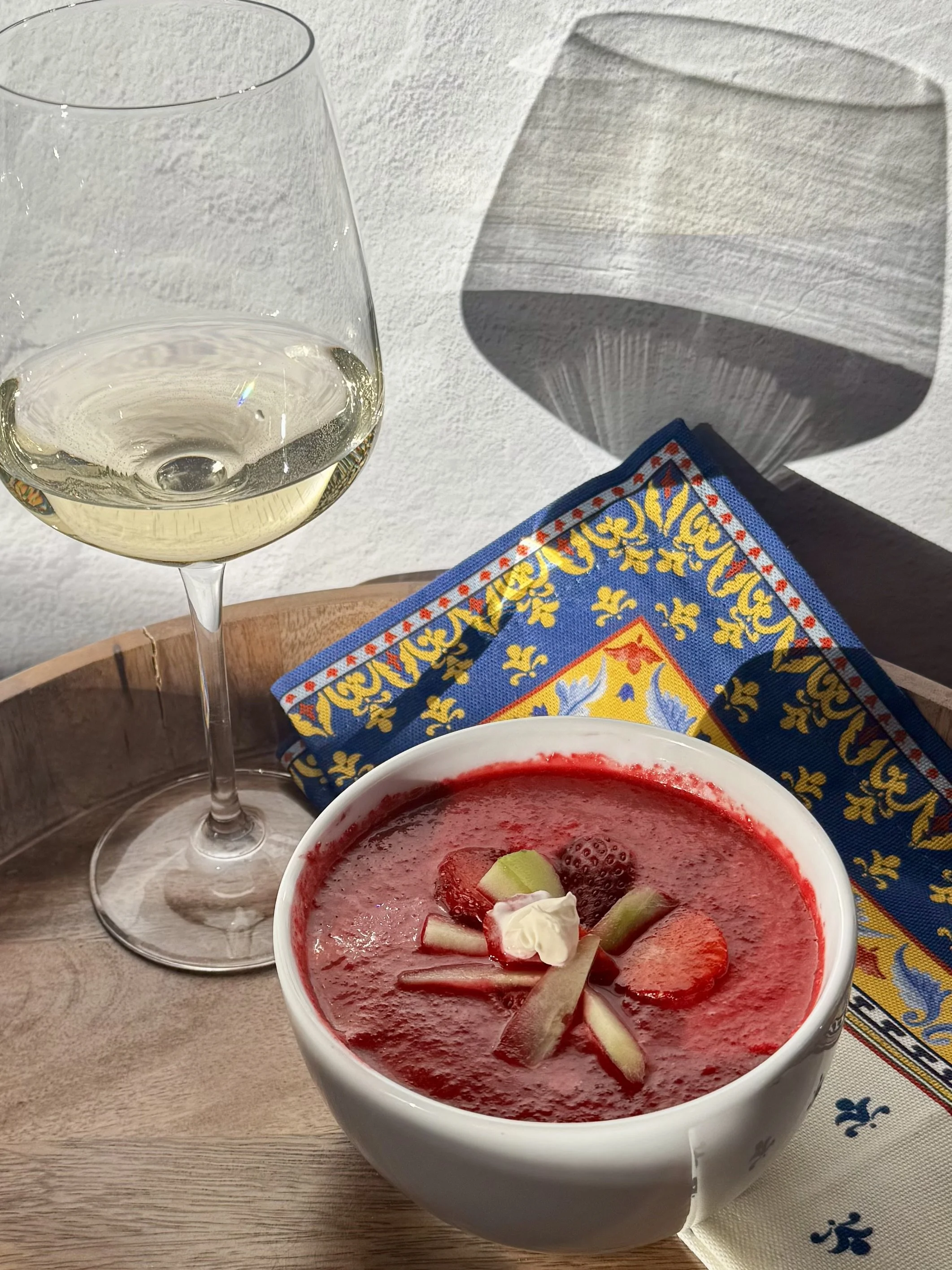 white wine in a glass on a wooden tray next to a white bowl with bright red gazpacho sitting on a cloth napkin