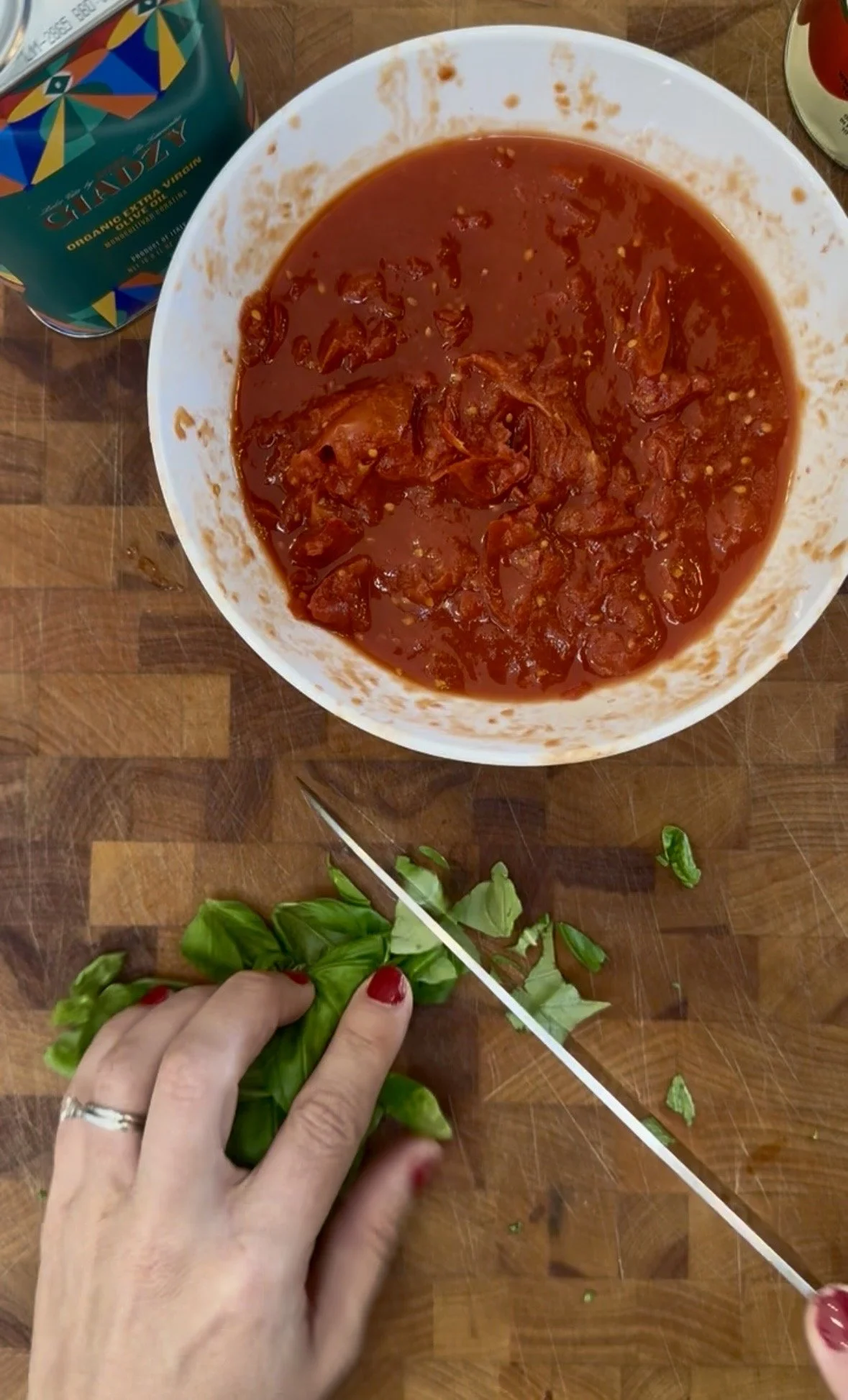 Hands chopping fresh basil on a butcher block with a bowl of crushed tomato sauce and Giadzy organic olive oil in the background