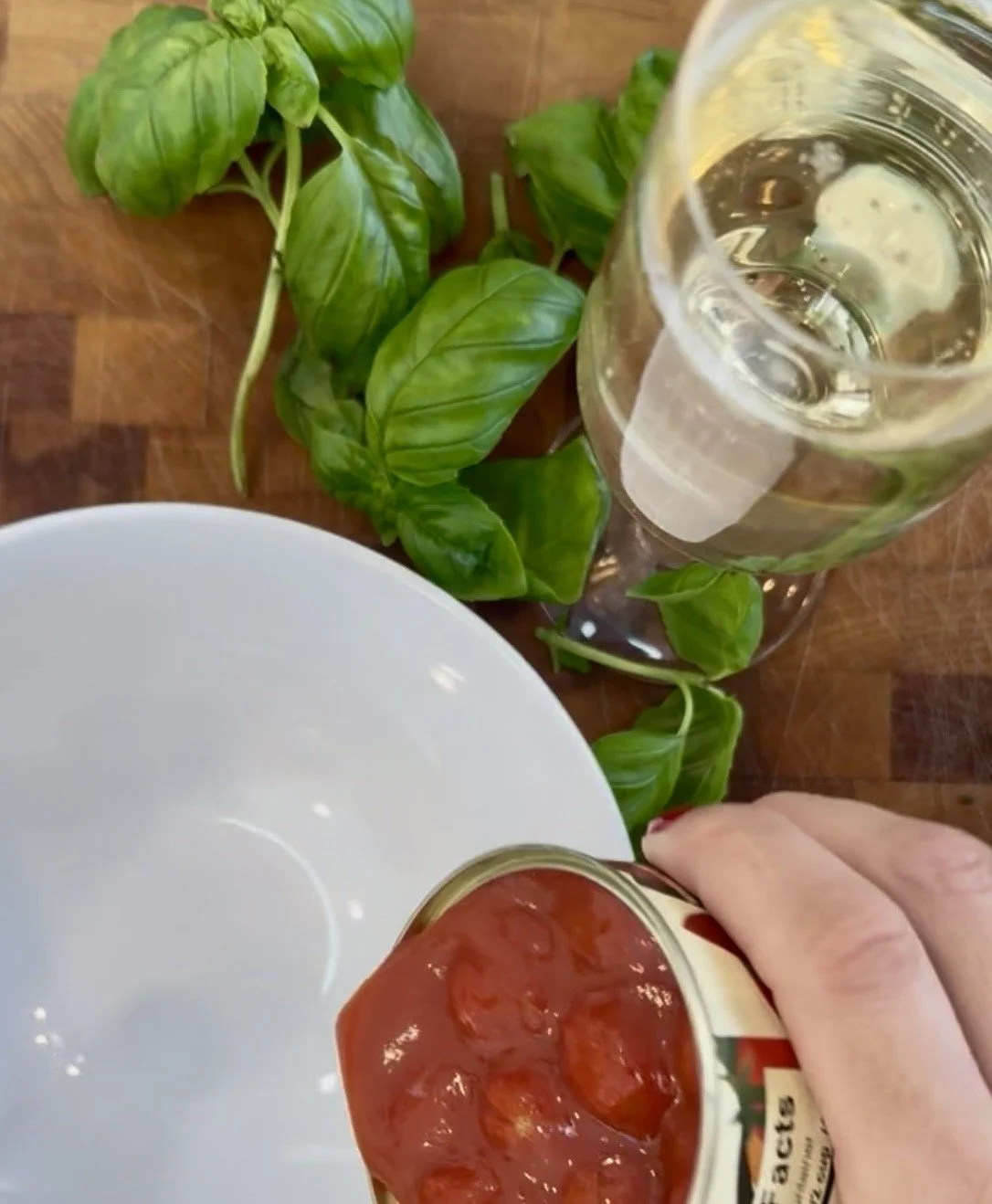Pouring Giadzy Pomodorino di Corbara tomatoes from the can into a white bowl with fresh basil and a glass of prosecco on a butcher block