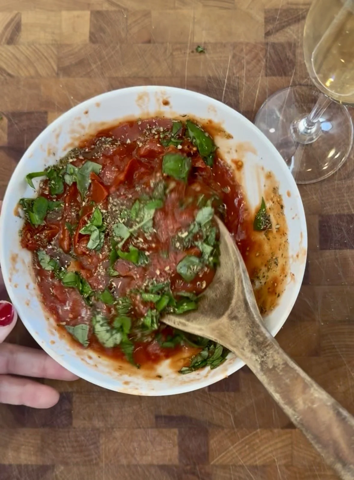 Finished no-cook hand-crushed tomato pizza sauce with fresh basil in a white bowl being stirred with a wooden spoon, prosecco glass in the background