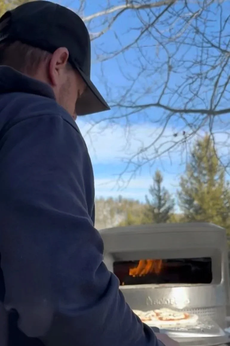 Man tending a Solo Stove Pie wood fired pizza oven outdoors with Montana pine trees and blue sky in the background