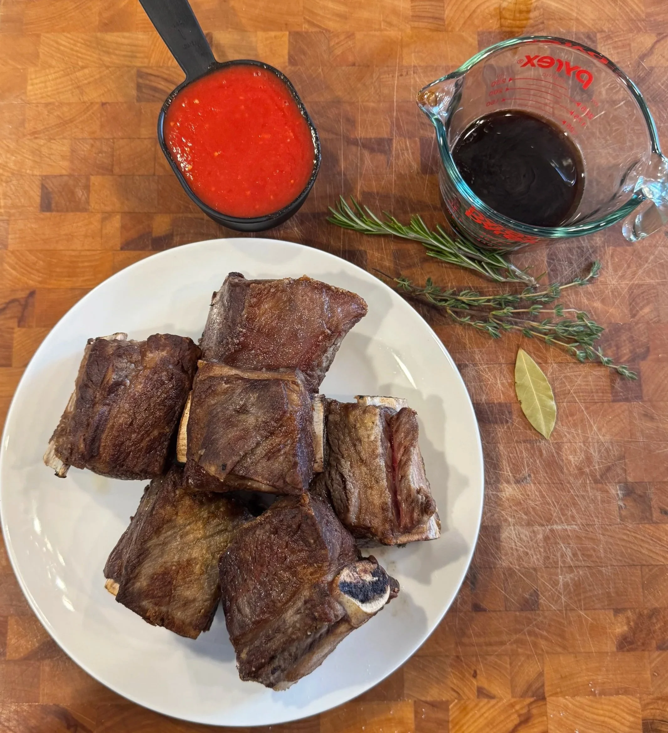 Ingredients for red wine braised short ribs including beef short ribs, tomatoes, red wine, rosemary, thyme, and bay leaf.