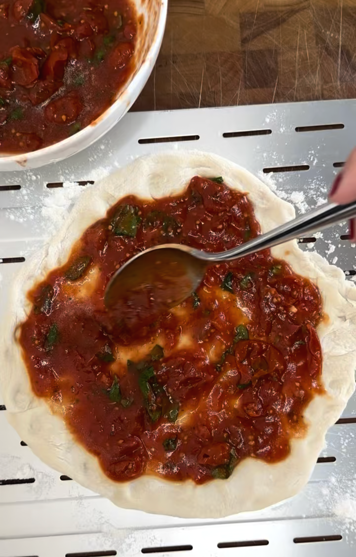 Spooning hand-crushed tomato sauce onto stretched homemade pizza dough on a perforated metal pizza peel dusted with flour