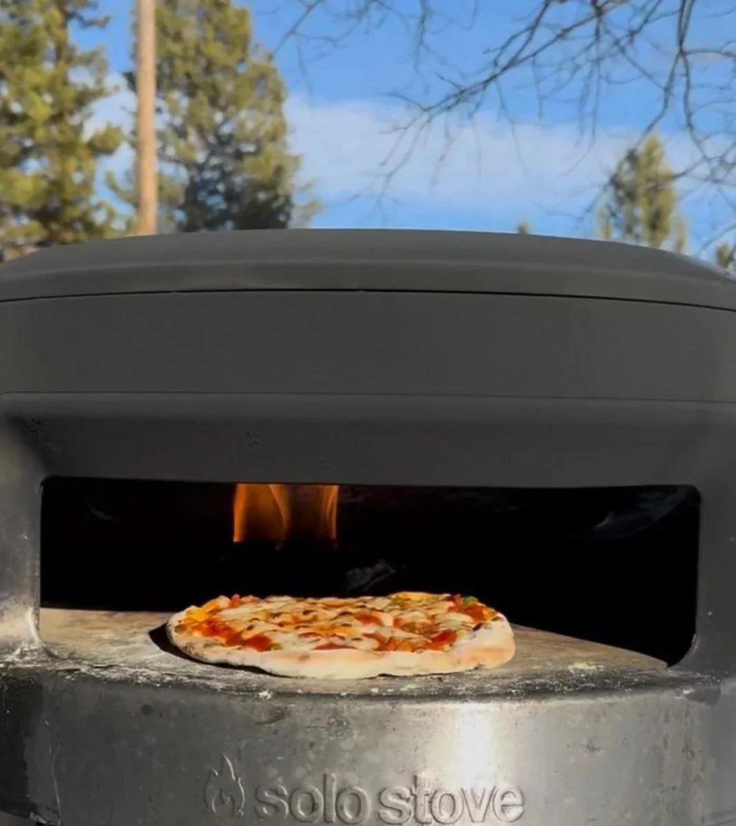 Solo Stove Pie wood fired pizza oven outdoors with a pizza cooking inside and Montana pine trees and blue sky in the background