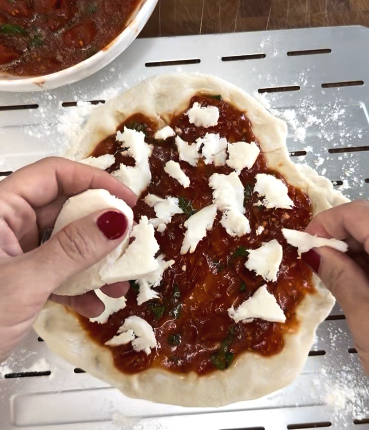 Hands tearing fresh mozzarella cheese over tomato-sauced homemade pizza dough on a metal pizza peel