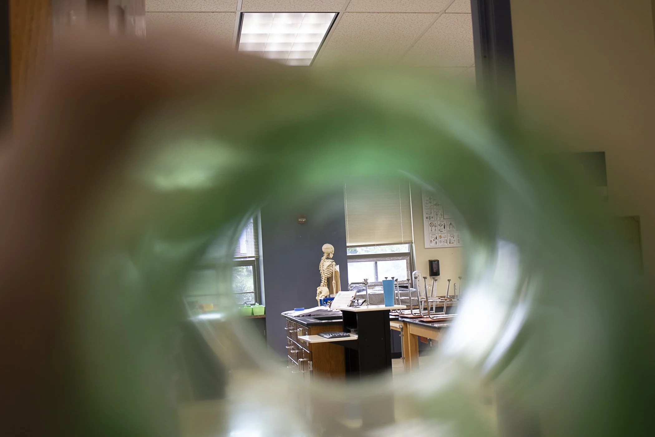 View of a classroom or laboratory seen through a blurry green object in the foreground, featuring an anatomical skeleton model, desks, chairs, and educational charts on the wall.