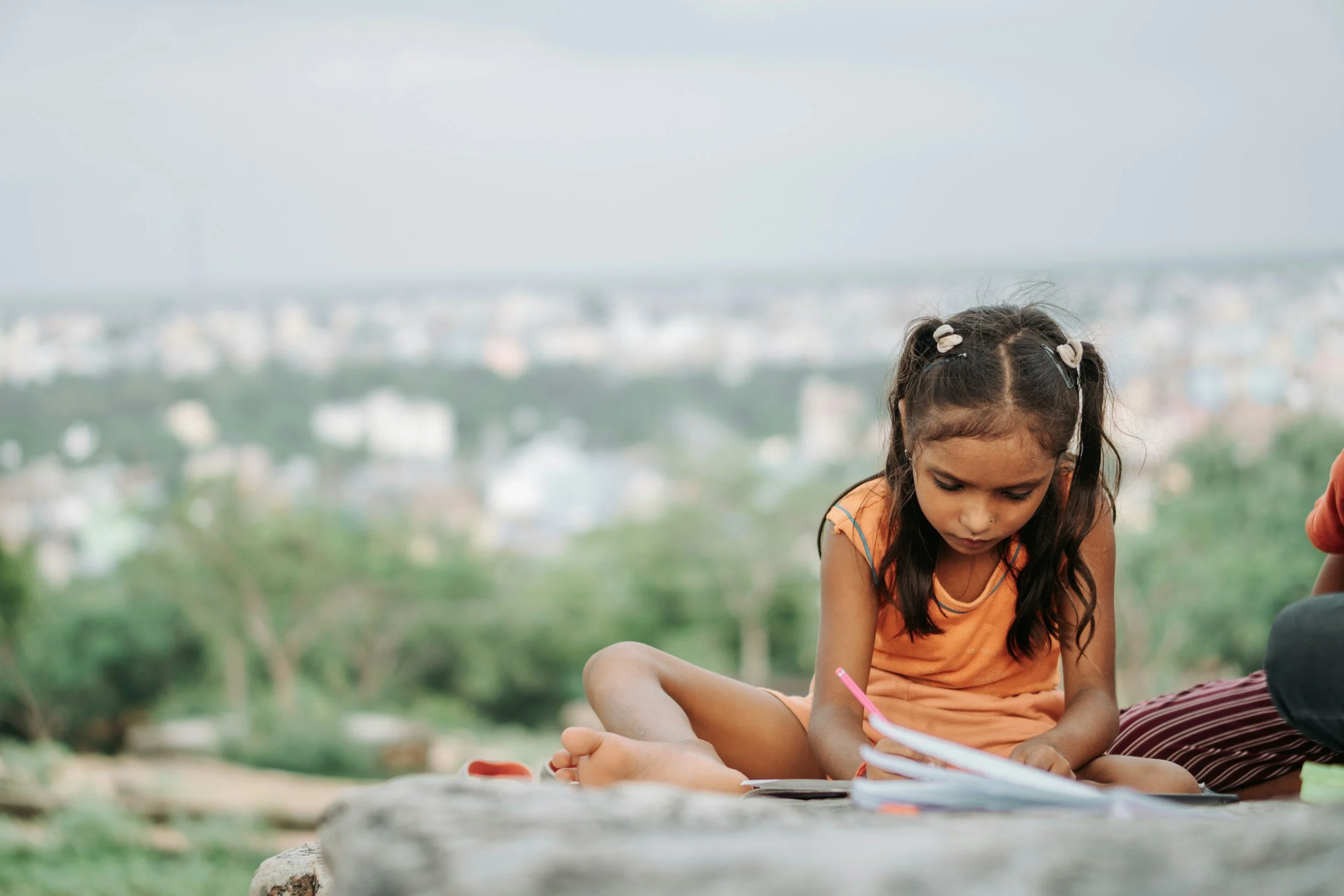 A young girl with pigtails sitting outdoors, writing in a notebook.