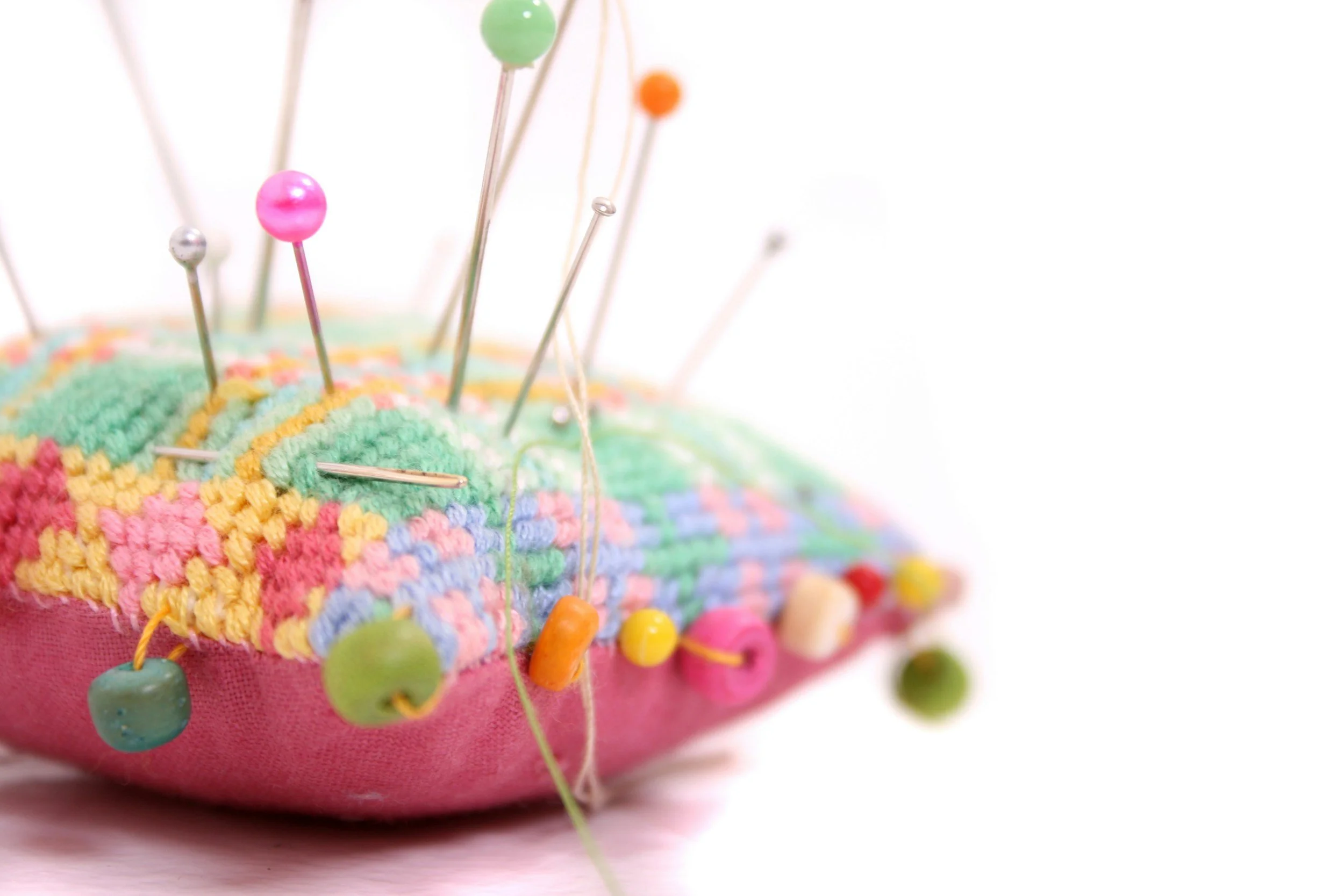 Close-up of a colorful embroidered pincushion with various pins and sewing needles stuck in it against a white background.