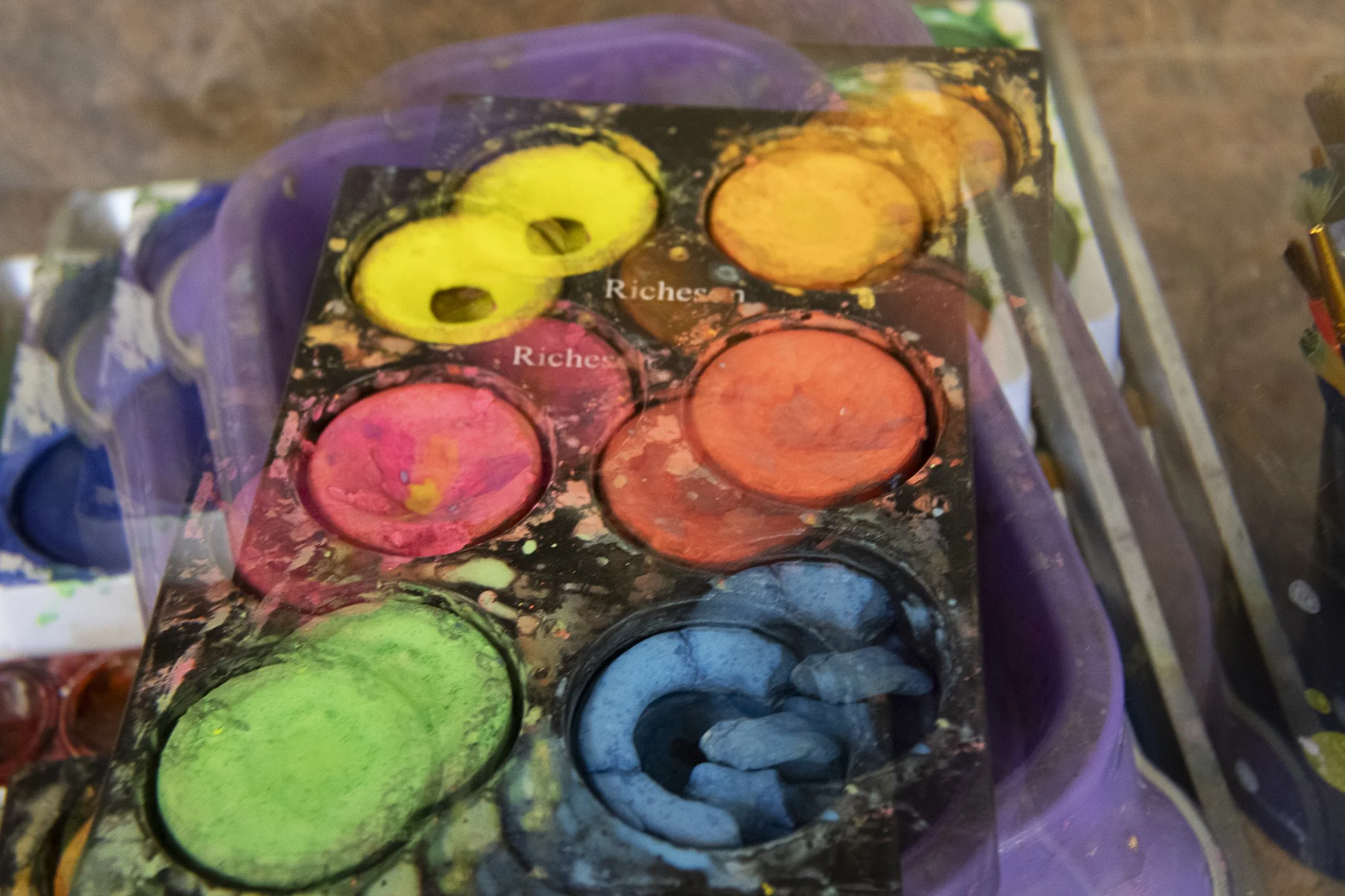 Colorful watercolor paint palette with various paint colors, some brushes, and a purple cloth underneath.